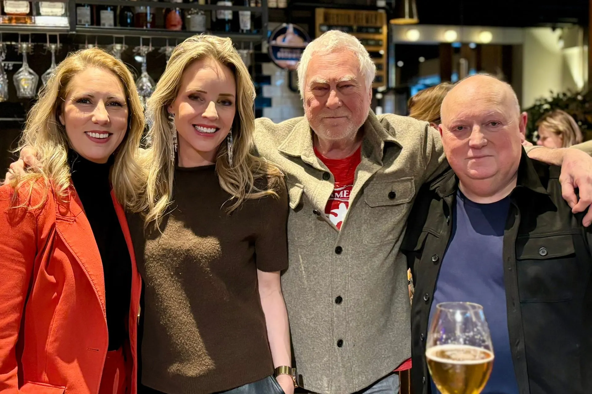 Gezellige borrelmomenten in een sfeervolle horecagelegenheid met een rijk assortiment dranken op de achtergrond. Vier personen staan dicht bij elkaar, gekleed in stijlvolle outfits, terwijl op de voorgrond een glas bier op tafel staat. De setting straalt warmte en gastvrijheid uit, ideaal voor netwerken, bedrijfsborrels en sociale evenementen. Perfect voorbeeld van een ontspannen vrijdagmiddagborrel in een moderne baromgeving met luxe interieur en gezellige ambiance. Omroep Brabant met Michelle de Wit en Cesar Zuiderwijk