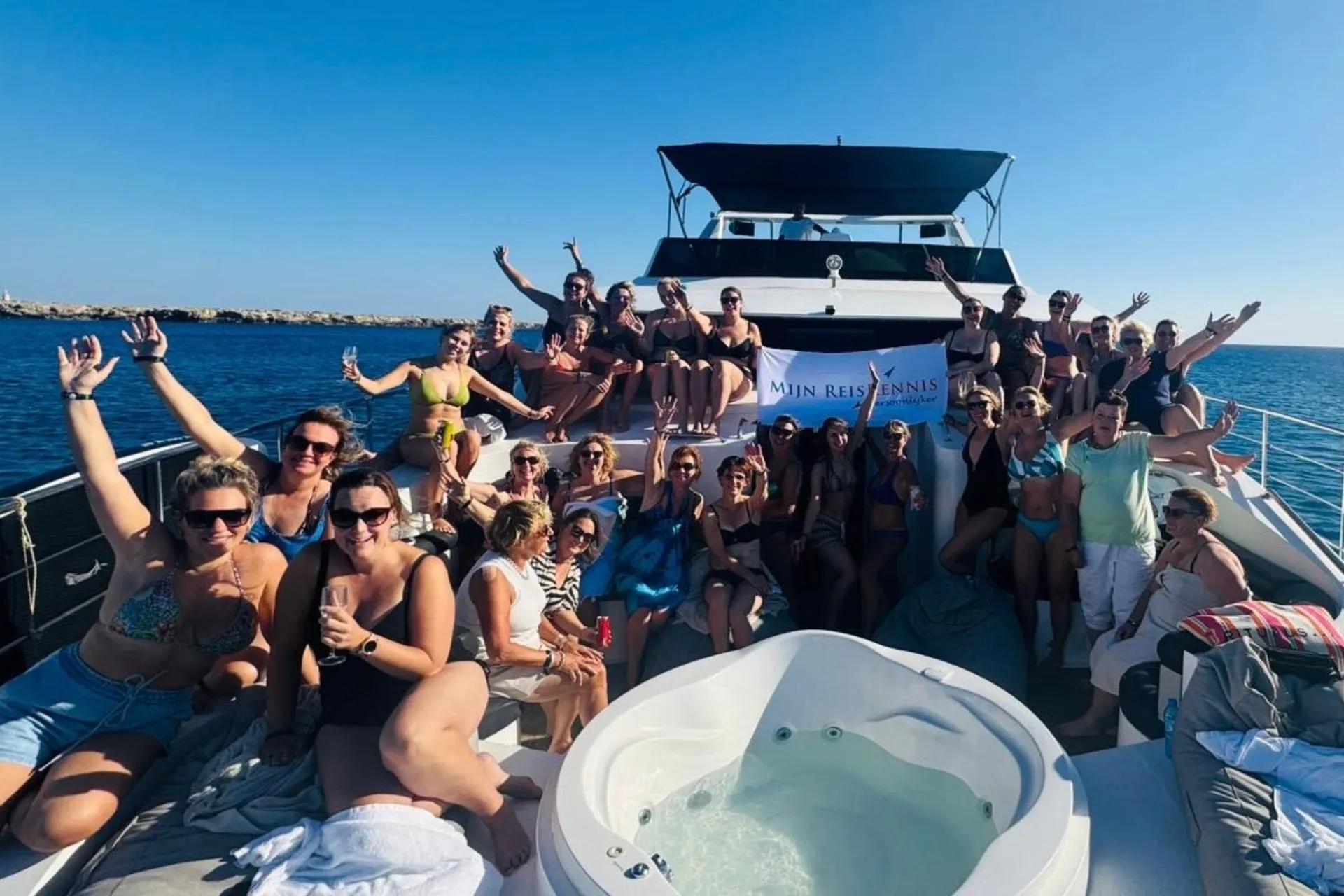 Grote groep mensen in badkleding op het dek van een boot met jacuzzi, voor een banner met de tekst “Mijn ReisKennis”. De zee en blauwe lucht op de achtergrond.