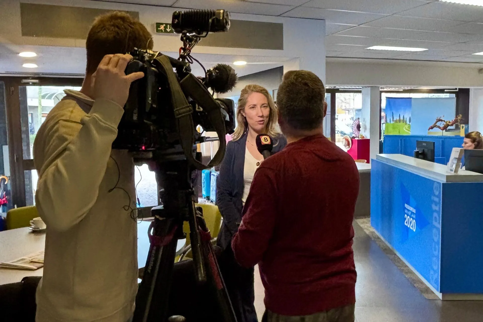 Michelle de Wit, eigenaresse van Travelcenter de Wit, wordt geïnterviewd door een journalist van Omroep Brabant in het reisbureau. Een cameraman filmt het gesprek terwijl de werkruimte en receptiebalie op de achtergrond zichtbaar zijn.