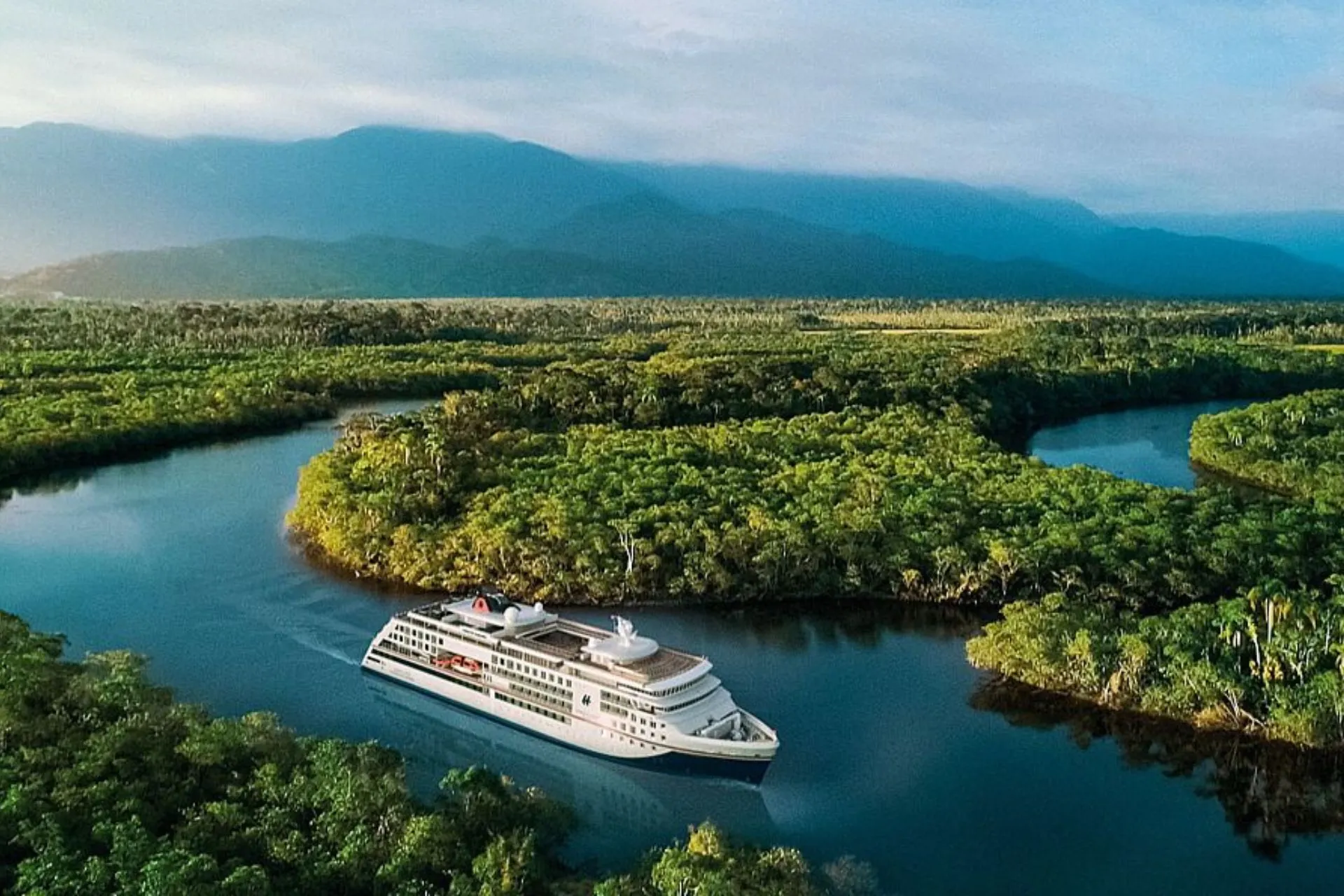 Luxe expeditiecruiseschip Hanseatic Spirit van Hapag Lloyd vaart door een kronkelende rivier omringd door dichtbegroeide jungle en groene bossen, met een bergachtig landschap op de achtergrond.