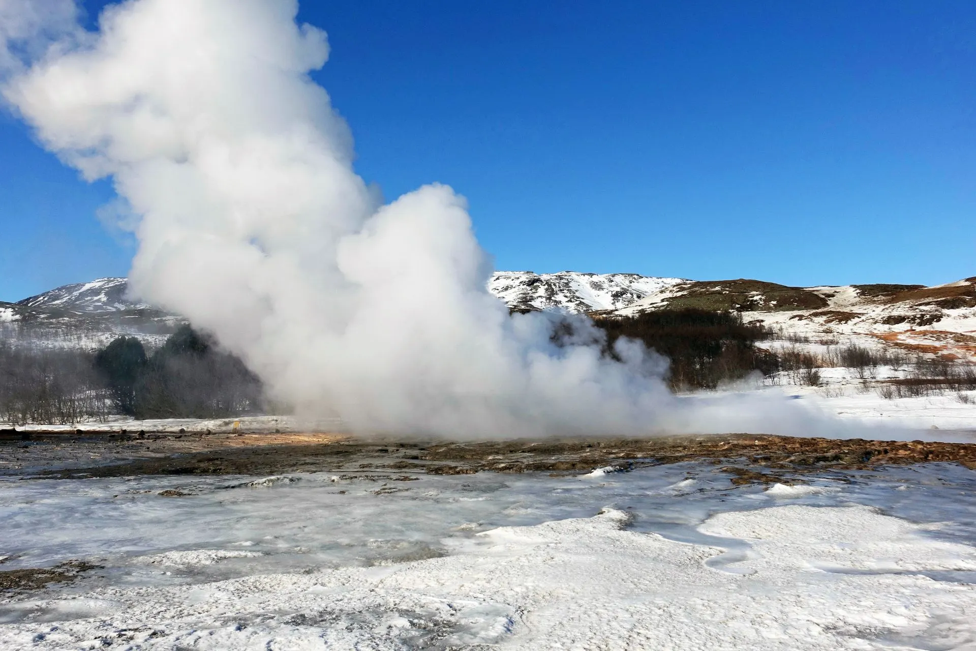 Een geiseruitbarsting van Strokkur in IJsland, met een wolk van stoom en besneeuwde bergen op de achtergrond onder een blauwe lucht.