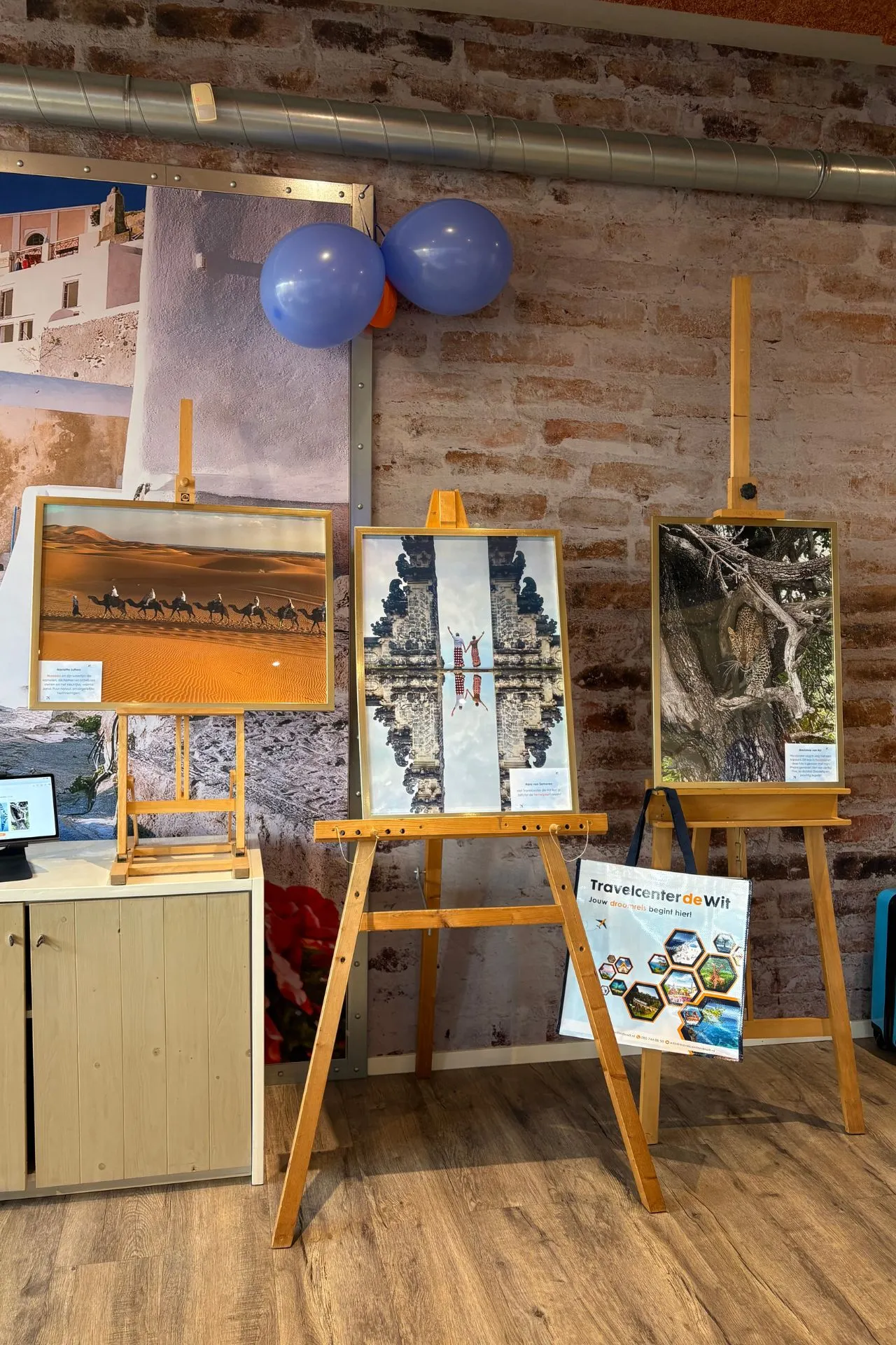 Drie genomineerde vakantiefoto’s op schildersezels tijdens het openingsfeest van Travelcenter de Wit Asten, met kleurrijke ballonnen en een Travelcenter de Wit-poster op de achtergrond.