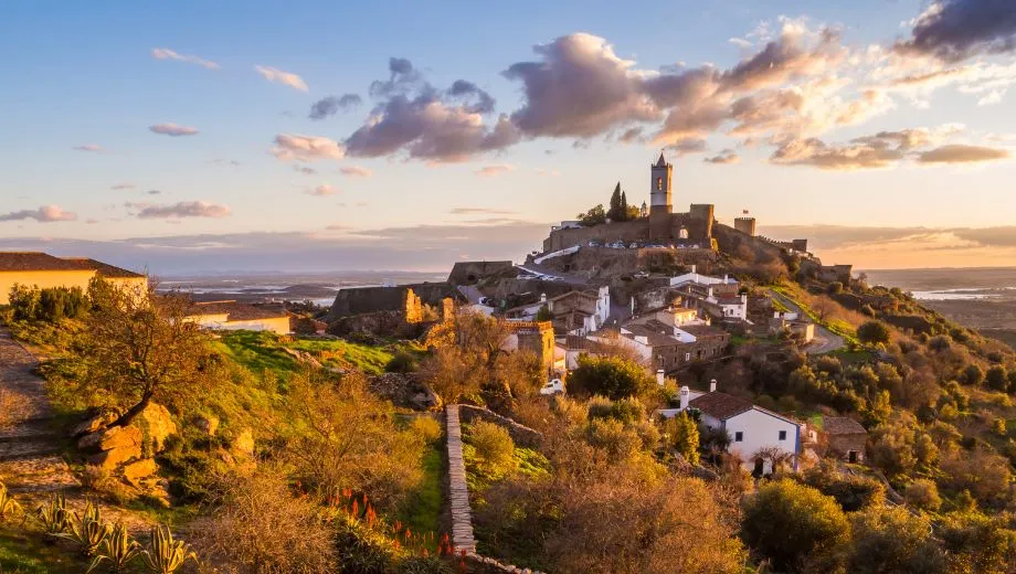 "Panoramisch uitzicht op het middeleeuwse dorp Monsaraz in Alentejo, Portugal, met een imposant kasteel op de heuveltop, witte huizen en kronkelende straatjes. De zonsondergang verlicht het landschap met gouden tinten, omringd door olijfbomen en groene valleien. Monsaraz is een populaire bestemming voor cultuur, geschiedenis en natuur in Alentejo. Ontdek authentieke Portugese charme, historische bezienswaardigheden en adembenemende uitzichten."