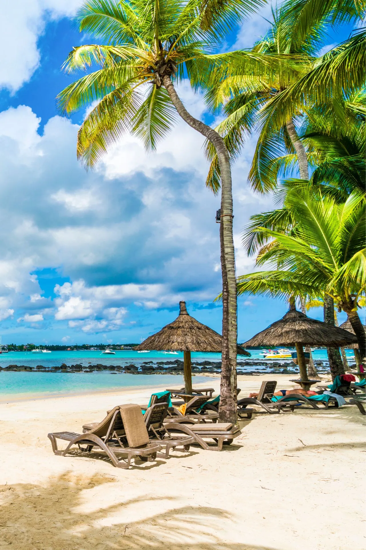 Een tropisch strand met witte zandstranden, rieten parasols en ligstoelen onder hoge palmbomen. Het turquoise water van de oceaan strekt zich uit naar de horizon, waar enkele boten dobberen. De lucht is deels bewolkt met blauwe stukken zichtbaar tussen de wolken. Een ontspannen vakantiesfeer hangt in de lucht.