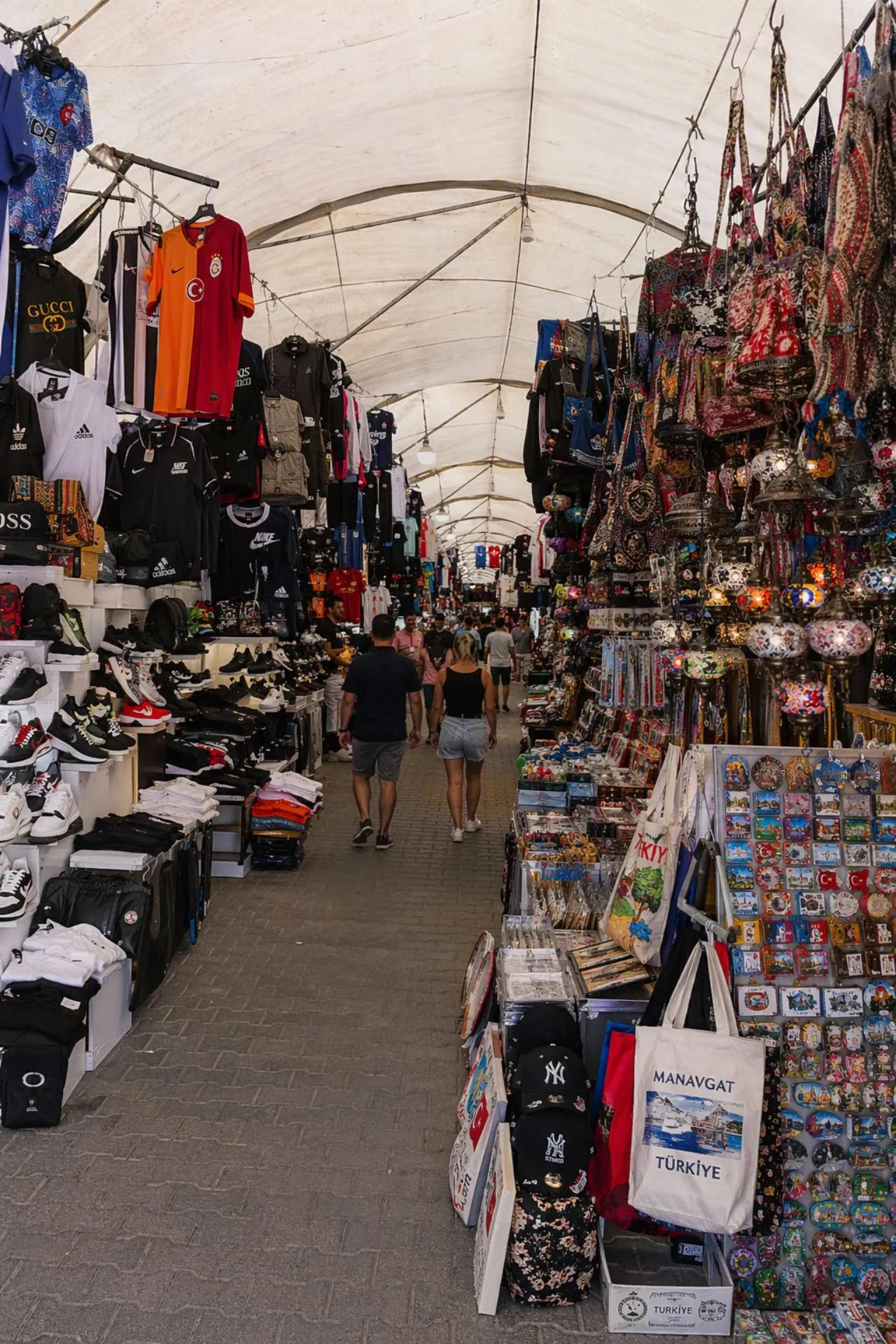 Markt van Manavgat in Turkije met kleurrijke kraampjes, kleding, tassen, souvenirs en lokale producten. Bezoekers wandelen door de overdekte markt terwijl winkeliers een breed aanbod presenteren. De populaire markt van Manavgat, vlak bij Side, is een geliefde excursie voor vakantiegangers die de Turkse cultuur en sfeer willen ervaren.