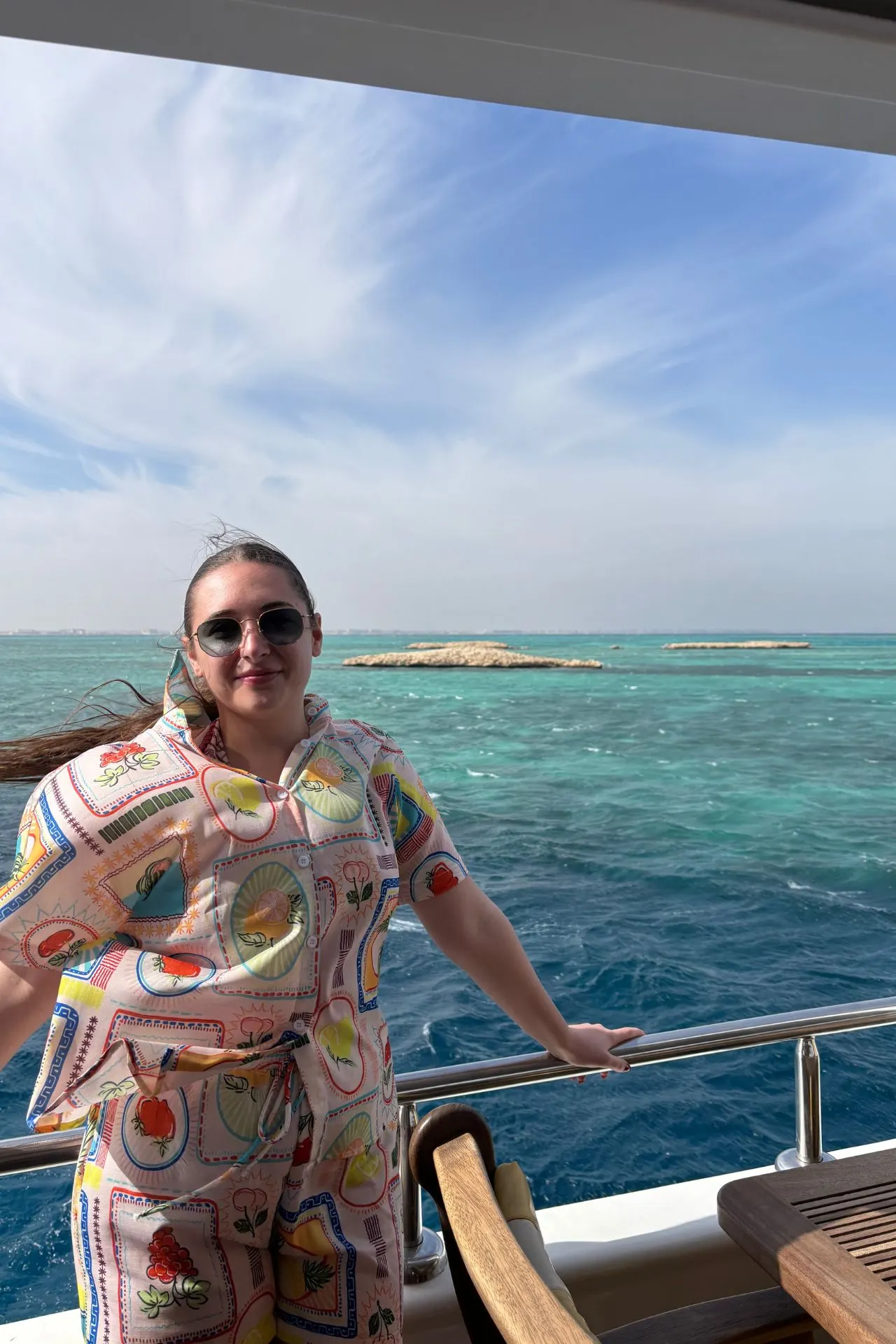 Persoon staat op het dek van een boot met uitzicht op helder turquoise water en kleine rotsachtige eilandjes in de verte. De lucht is blauw met lichte, uitwaaierende wolken, wat een zonnige en ontspannen sfeer creëert. De persoon draagt een kleurrijke outfit met een patroon van fruit en decoratieve elementen, terwijl een houten stoel op de voorgrond zichtbaar is. Dit beeld benadrukt luxe en ontspanning tijdens een boottocht, ideaal voor promotie van strandvakanties, cruises en avontuurlijke reizen in Egypte.