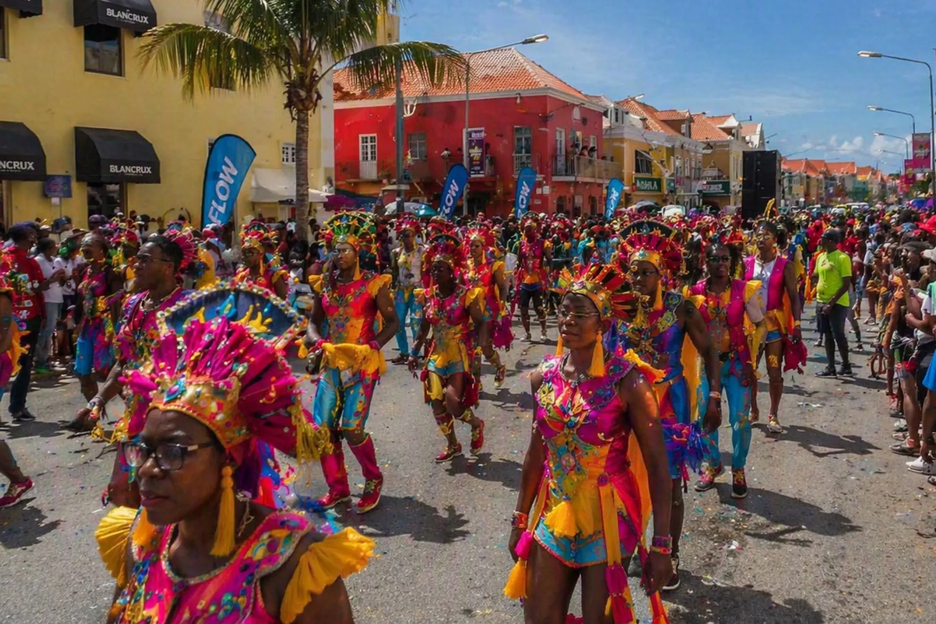 Grote carnavalsoptocht in Willemstad op Curaçao met dansers in opvallende kostuums en veel publiek langs de straat. De kleurrijke gebouwen maken de feestelijke sfeer compleet.