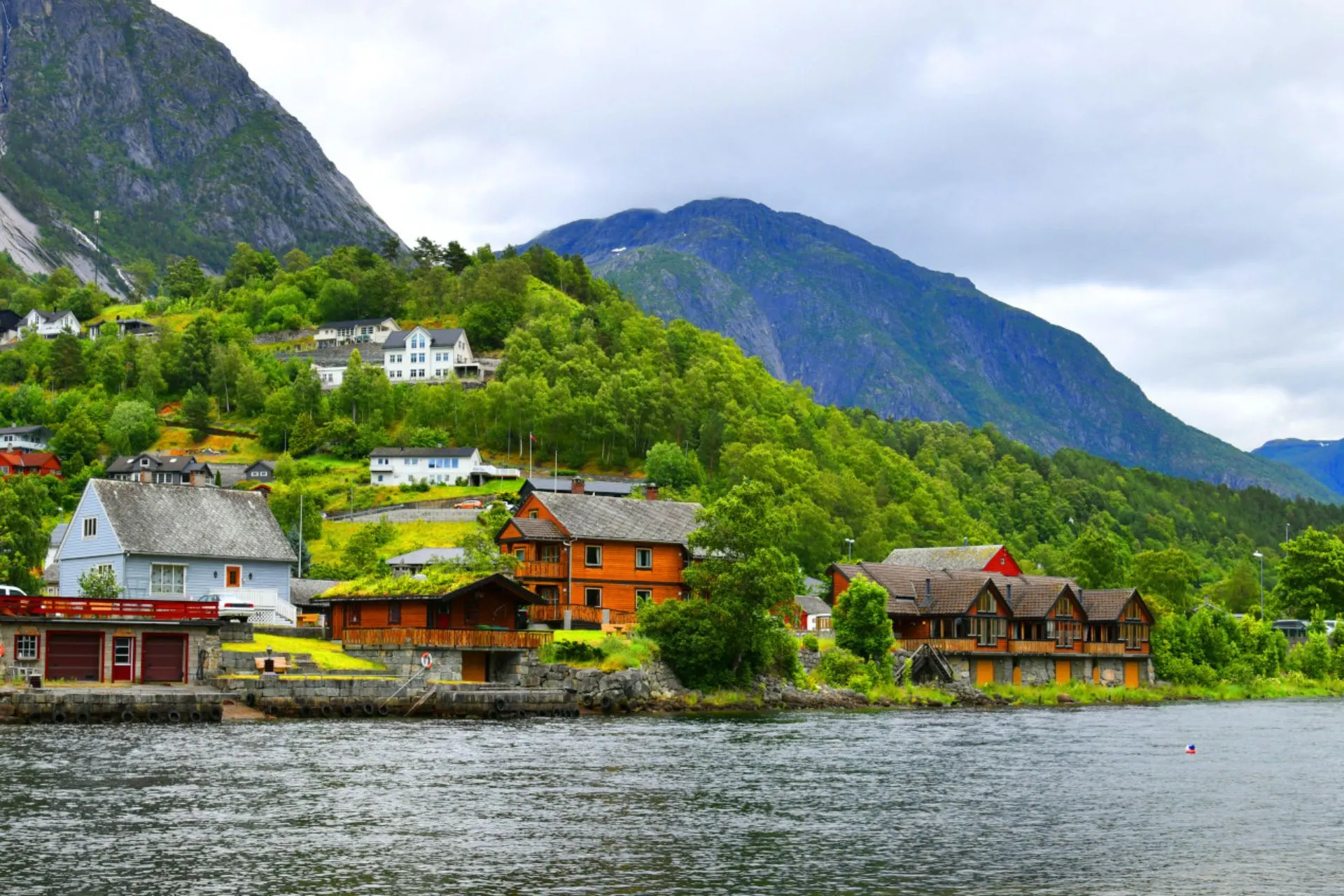 Kleurrijke Noorse huisjes in Eidfjord met groene heuvels en bergachtige achtergrond onder een halfbewolkte lucht.
