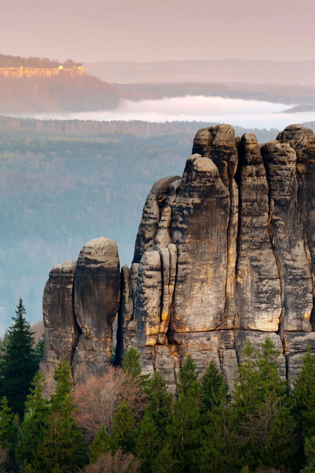 "Adembenemend uitzicht op Saksisch Zwitserland in Duitsland, met een vallei bedekt door dichte mist en kleurrijke velden op de achtergrond. Dit nationale park staat bekend om zijn spectaculaire rotsformaties, wandelroutes en schilderachtige landschappen. Perfect voor natuurliefhebbers en fotografen, ontdek de unieke combinatie van bossen, bergen en pittoreske dorpen in het hart van Oost-Duitsland."