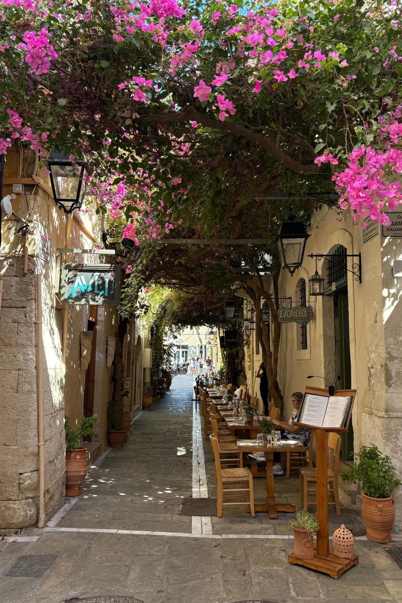 Sfeervol straatje in Rethymnon met een overkapping van felroze bougainvillea en gezellige terrastafeltjes langs de muur.