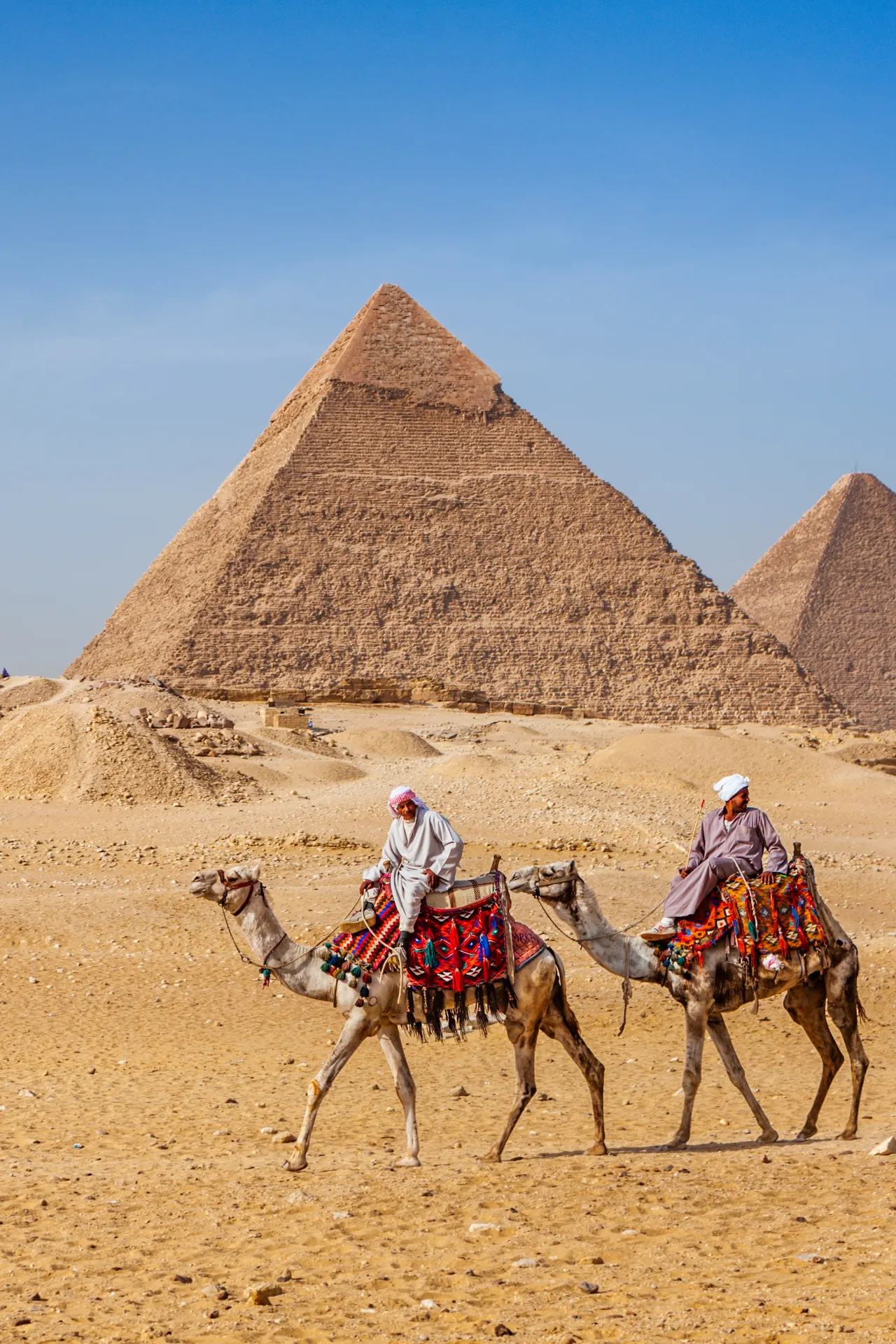 Twee mannen rijden op kamelen in traditionele Egyptische kledij met piramides op de achtergrond, onder een heldere blauwe lucht in een woestijnlandschap.