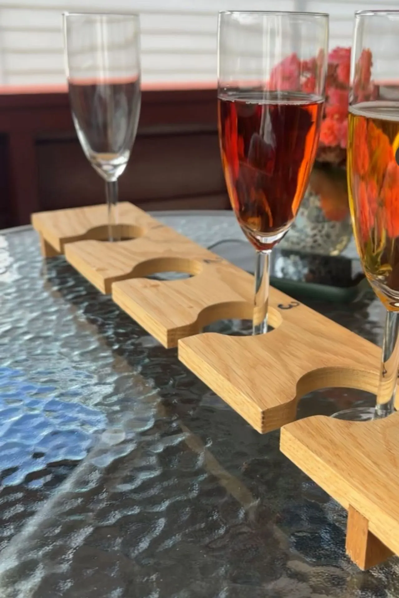 Drie champagneglazen met verschillende dranken in een houten houder op een glazen tafel. Op de achtergrond een vaas met roze bloemen en een witte wand, met zonlicht dat schaduwen werpt.