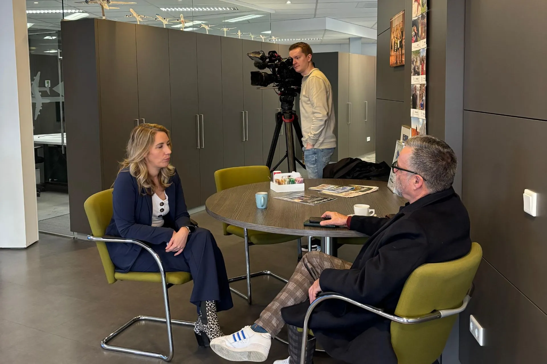 Michelle de Wit, eigenaresse van Travelcenter de Wit, zit aan een tafel in gesprek met een journalist van Omroep Brabant. Op de achtergrond staat een cameraman die het interview filmt. De setting is een moderne kantoorruimte met een informele sfeer.