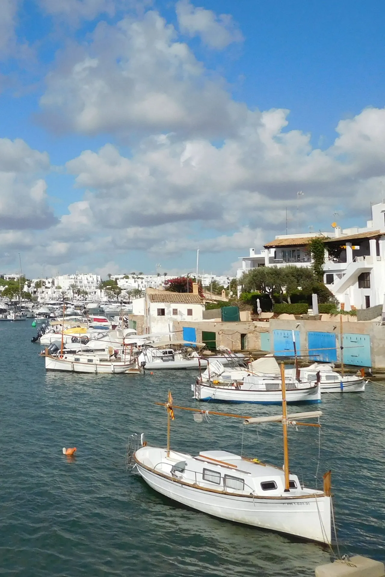 Haven van Cala d’Or op Mallorca met traditionele vissersboten en witte mediterrane huizen langs het water, onder een blauwe lucht met wolken. Deze idyllische kustplaats in het oosten van Mallorca staat bekend om zijn schilderachtige baaitjes, gezellige autovrije centrum en helderblauw water. Perfecte bestemming voor een zonvakantie, boottocht of ontspannen wandeling langs de haven.
