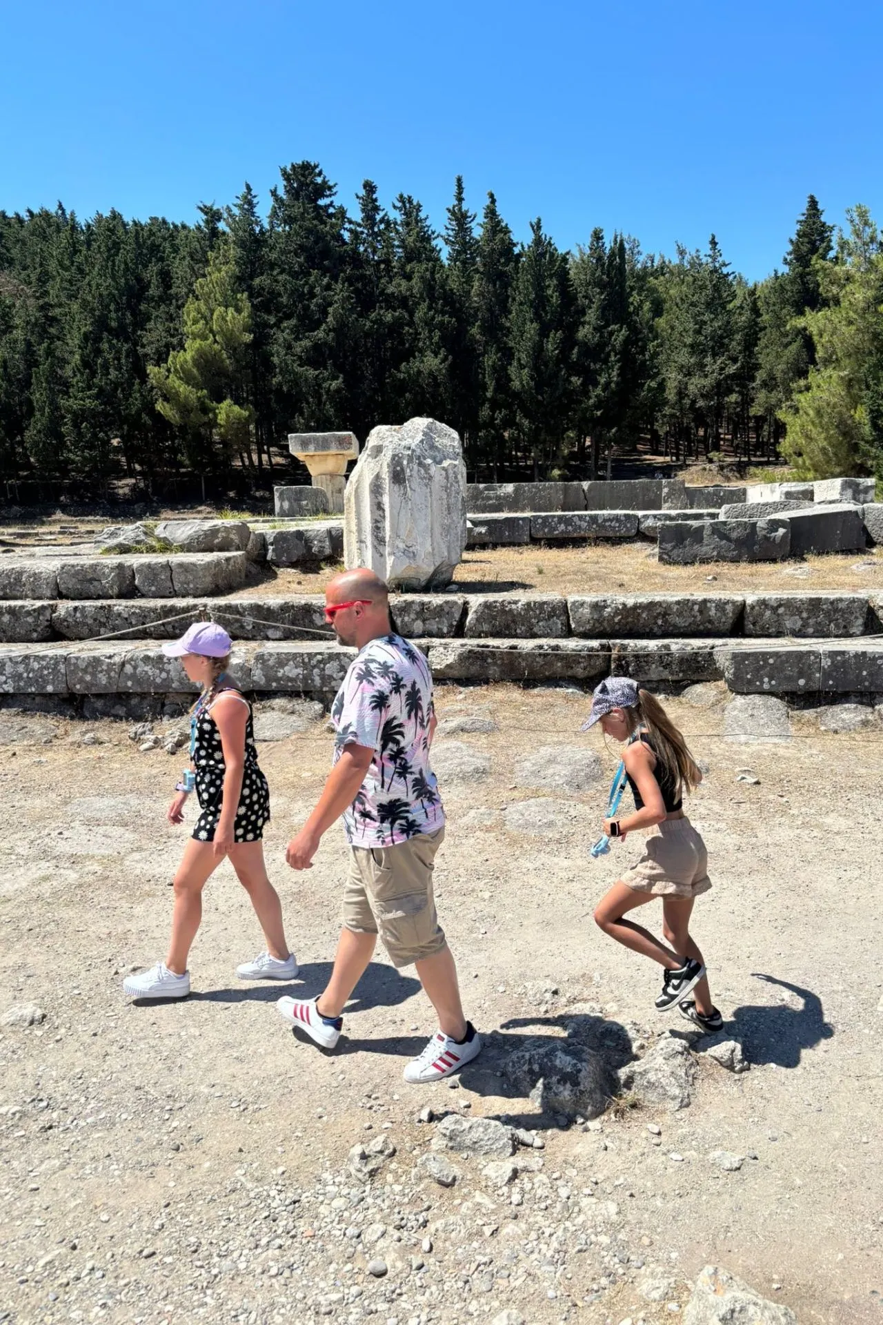 Ouders met kind lopen langs oude stenen fundamenten en zuilfragmenten van het Asklepion op Kos, omringd door dennenbomen en een blauwe lucht