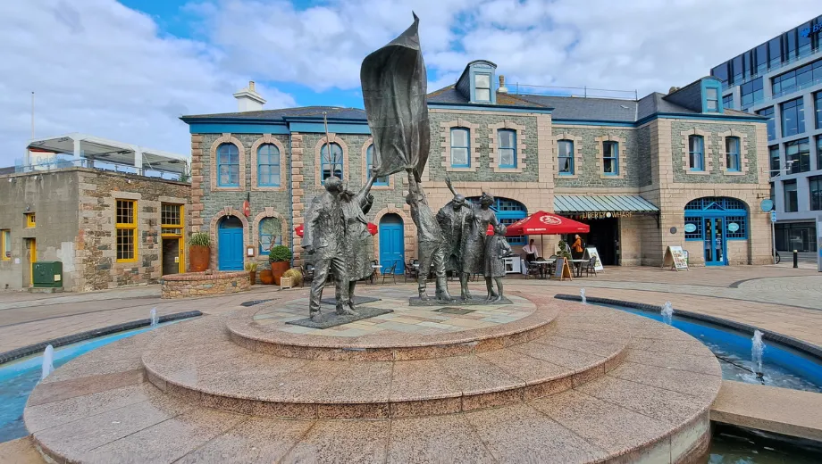Historisch plein met een opvallend bronzen standbeeld van drie figuren die een vlag omhoog houden, geplaatst op een ronde fontein met waterstralen. Op de achtergrond staan traditionele stenen gebouwen met blauwe deuren en ramen, naast moderne architectuur en een terras met rode parasol. Deze locatie is een populaire bezienswaardigheid en fotospot, ideaal voor toeristen die cultuur, geschiedenis en architectuur willen ontdekken in een levendige stadssfeer.