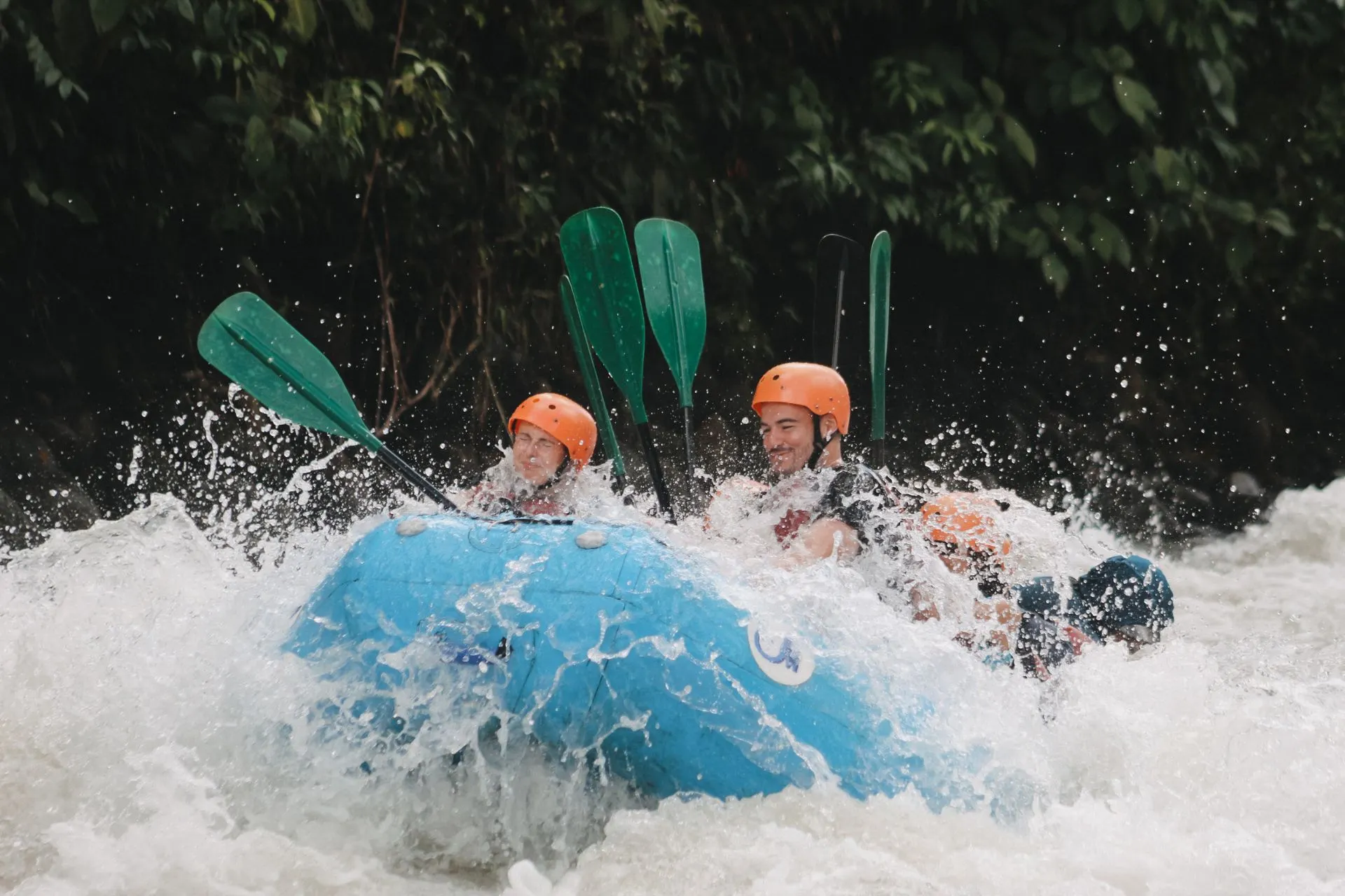Twee personen in een blauwe rubberboot raften door wilde stromingen, met opspattend water en groene natuur op de achtergrond.