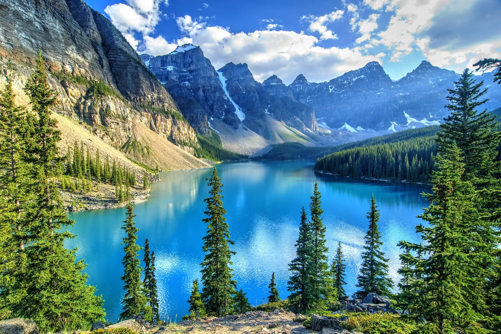 Adembenemend uitzicht op Moraine Lake in de Rocky Mountains, omringd door dichte dennenbossen en imposante bergtoppen met sneeuw. Het kristalheldere turquoise water weerspiegelt de steile rotswanden en groene bomen, wat een serene en natuurlijke sfeer creëert. Perfecte bestemming voor outdooractiviteiten zoals wandelen, kajakken en fotografie. Ideaal voor reizigers die avontuur, natuurbeleving en het ontdekken van de ongerepte schoonheid van de Canadese Rockies zoeken.