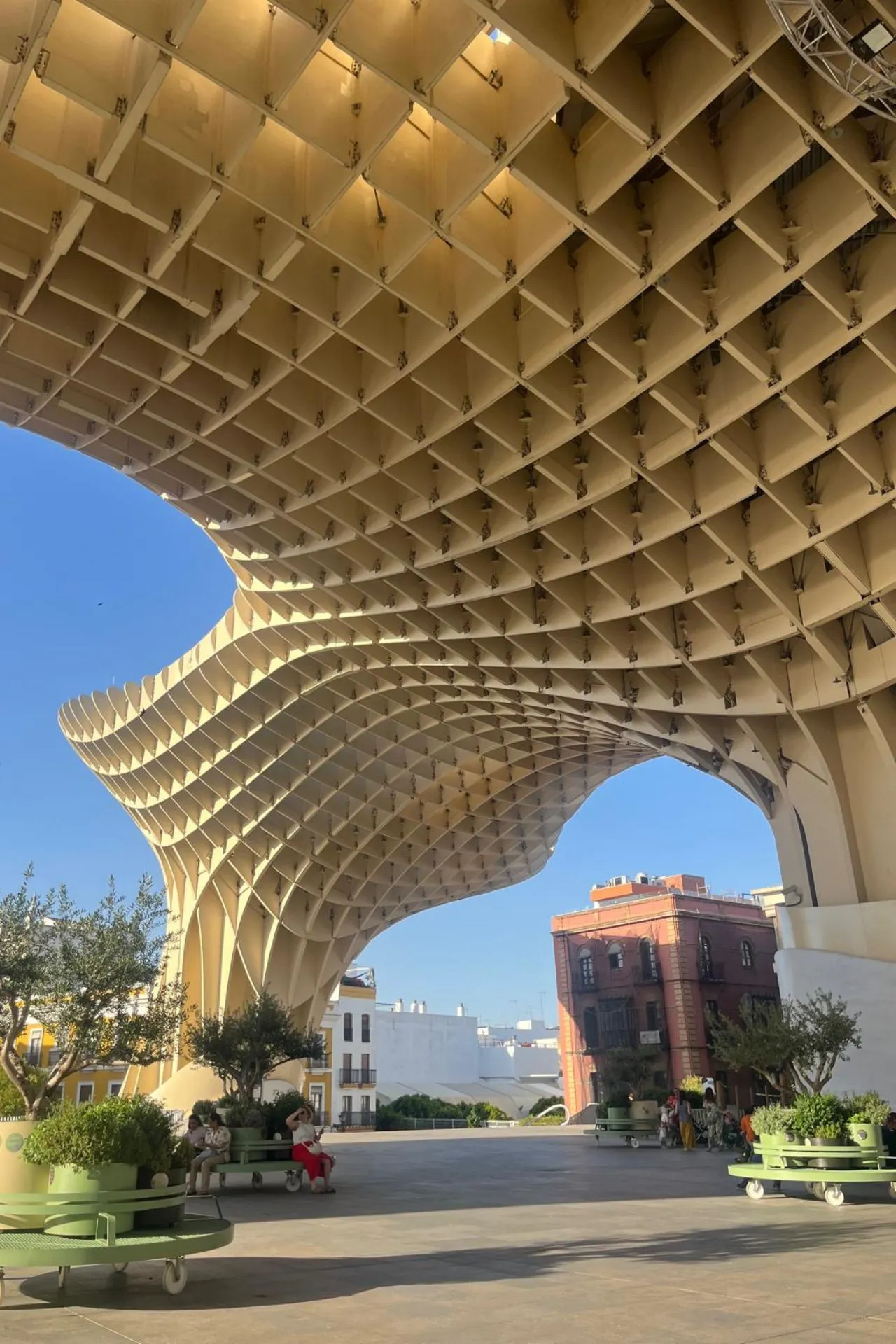 Onderzijde van de Metropol Parasol in Sevilla, met zicht op de gebogen houten panelen en schaduwrijke zitplekken met plantenbakken. Enkele mensen zitten op bankjes onder de constructie.