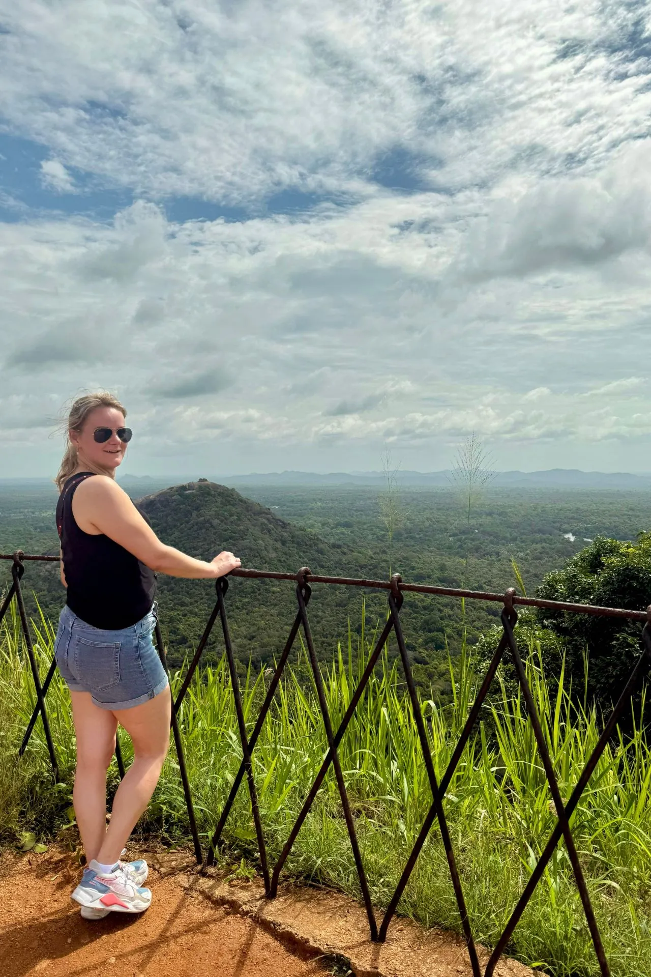 Uitzicht op de imposante Sigiriya-rots, gezien vanaf de tuinen aan de voet van het complex. De rots, met zijn opvallende verticale strepen in oranje- en grijstinten, rijst hoog boven het weelderige groene landschap uit. In de rotswand is een nis zichtbaar waarin de beroemde fresco's van de hemelse maagden zich bevinden. De symmetrische paden in de voorgrond leiden bezoekers door de historische water- en rotstuinen naar deze UNESCO-werelderfgoedlocatie.