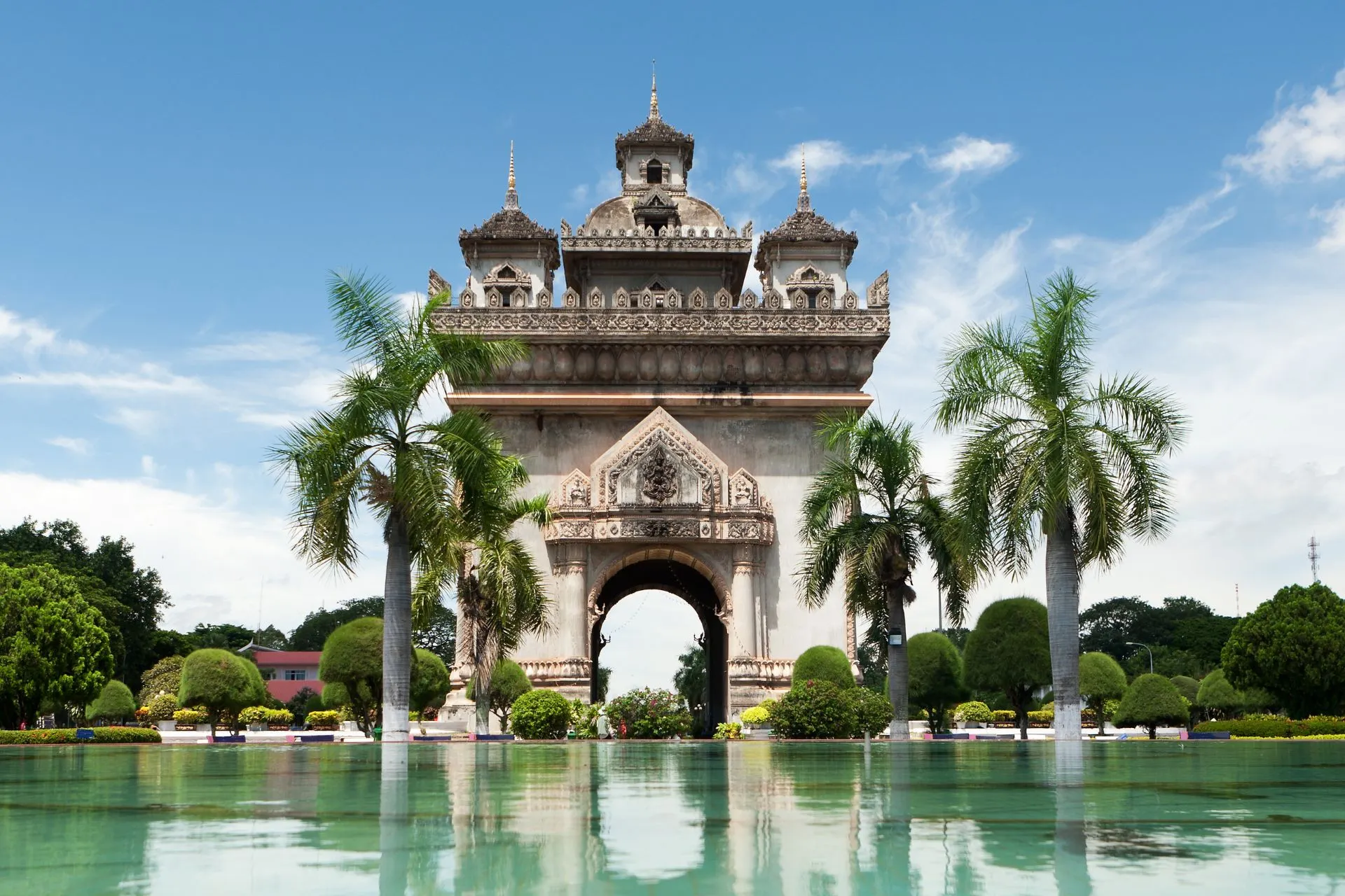 Decoratieve monumentale stadspoort met sierlijke details, omringd door palmbomen en een reflectie in een vijver.
