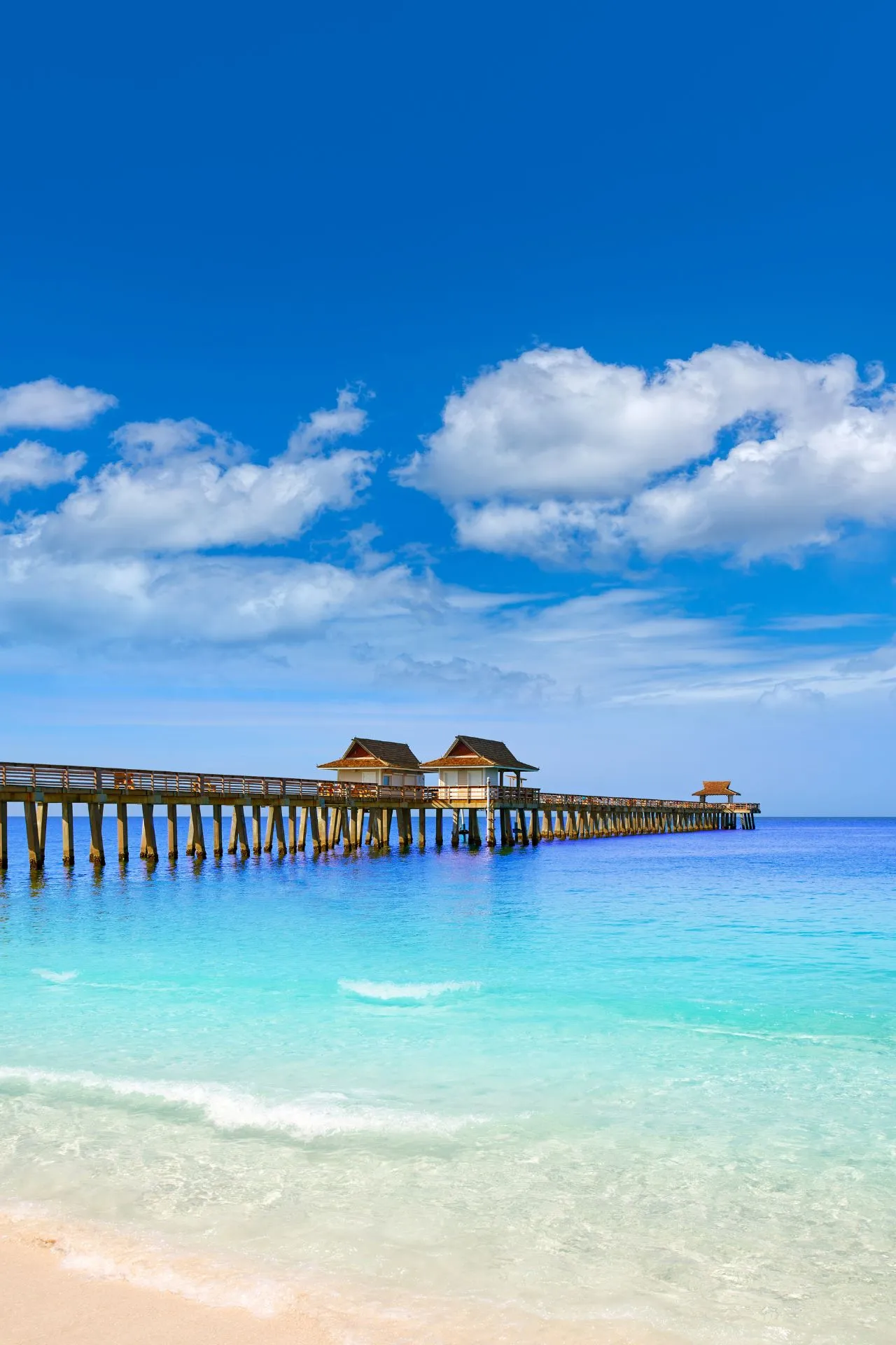 Pier in Naples, Florida, die zich uitstrekt over helderblauw water met wit zand op de voorgrond en een strakblauwe lucht erboven.