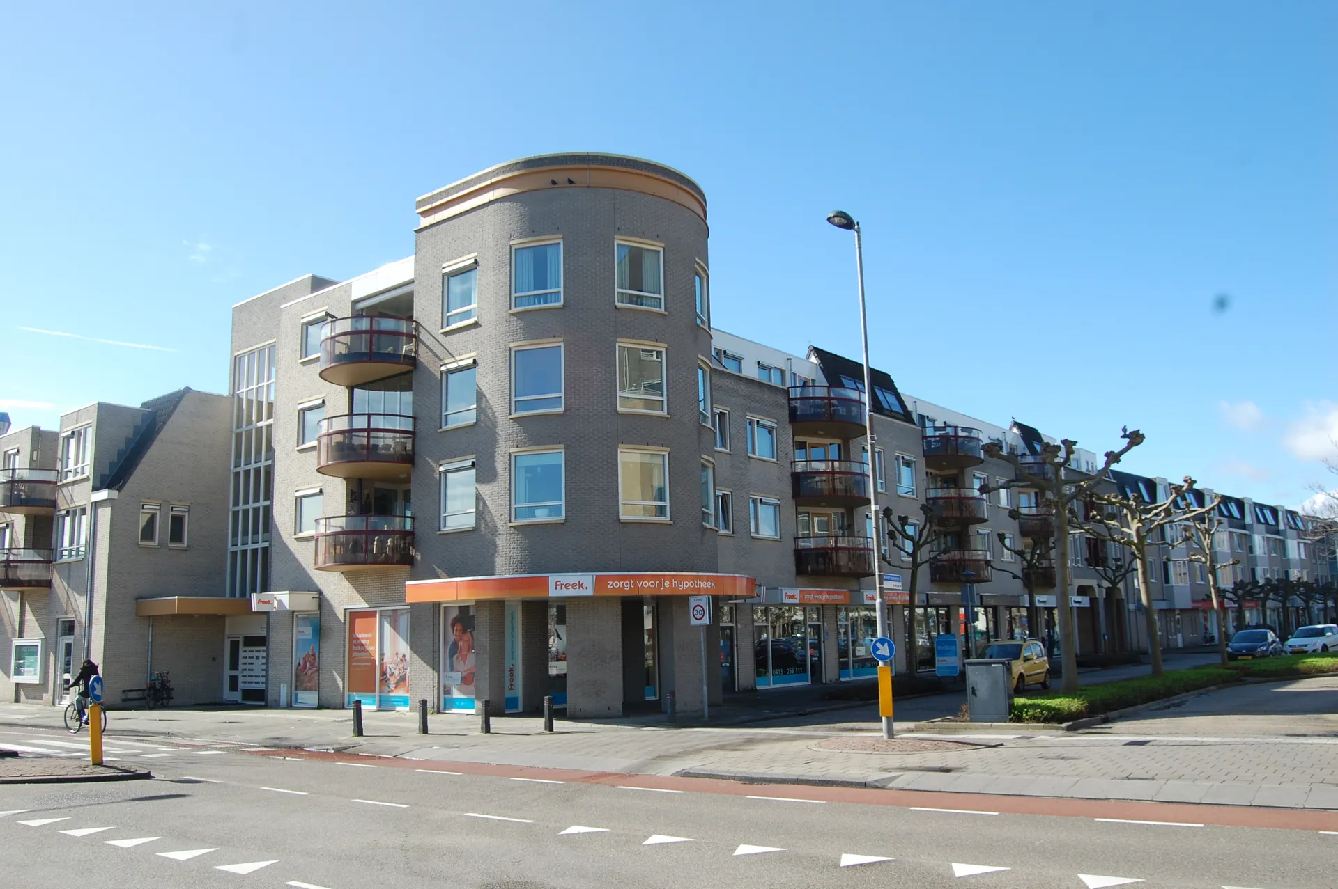  "Straatbeeld van een modern hoekgebouw met meerdere verdiepingen, grote ramen en ronde balkons, gelegen aan een kruispunt in een stedelijke omgeving. Op de begane grond bevinden zich commerciële ruimtes met oranje gevelstroken en etalages, ideaal voor winkels of kantoren. Het gebouw heeft een eigentijdse architectuur met lichte bakstenen en een strakke uitstraling. Perfecte locatie voor zakelijke dienstverlening, retail en stedelijke ontwikkeling in een goed bereikbare wijk."
