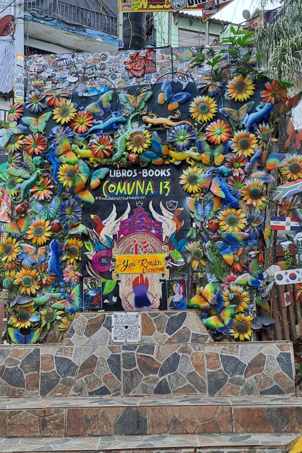 Kleurrijke muurschildering en kunstinstallatie in Comuna 13, versierd met bloemen, hagedissen en felgekleurde patronen, met tekst “Libros - Books Comuna 13”.