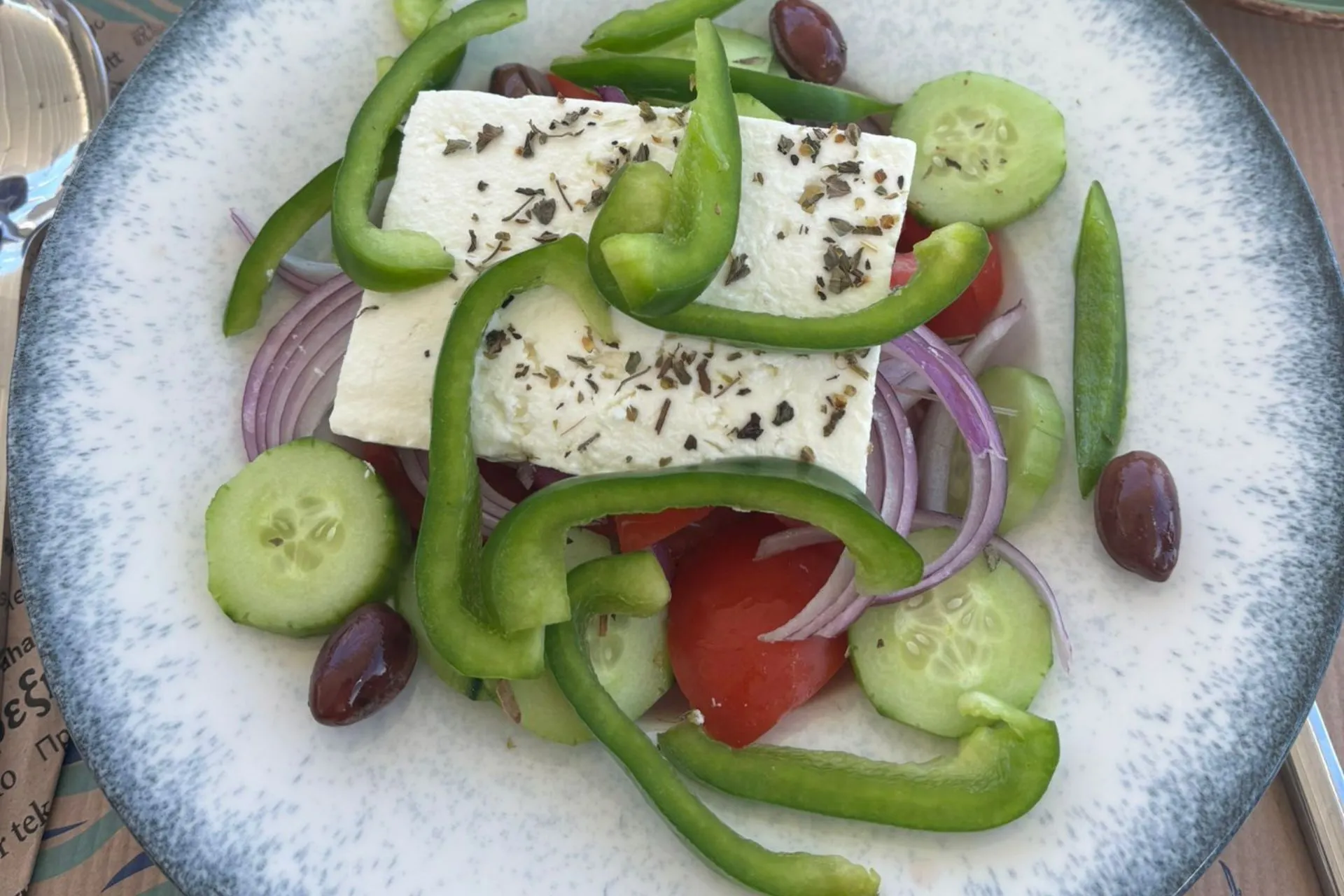 Bord met Griekse salade bestaande uit feta, komkommer, tomaat, rode ui, groene paprika en olijven, geserveerd op een tafel.