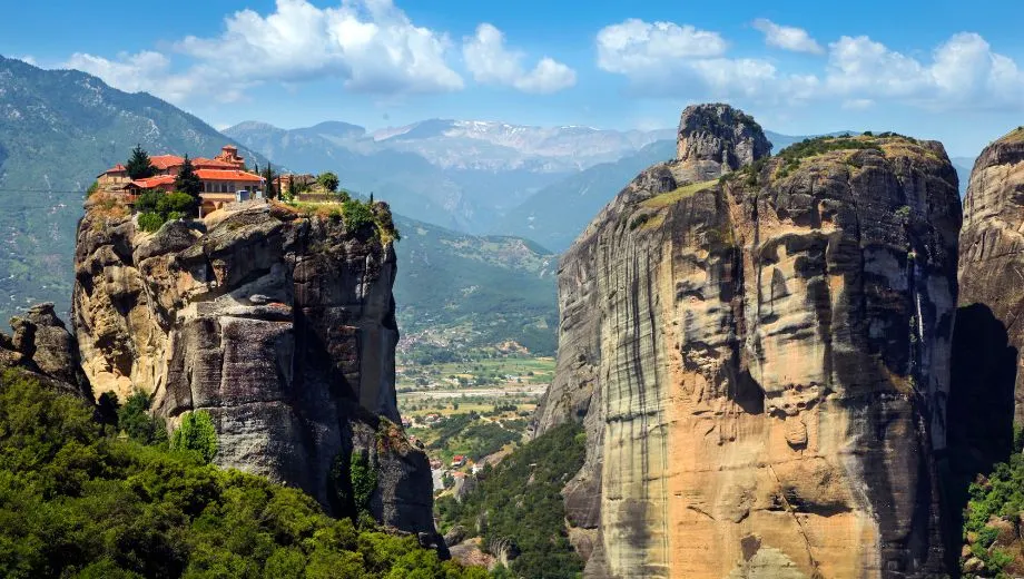 "Spectaculair uitzicht op Meteora in Griekenland, met een klooster op een steile rotsformatie en een groene vallei omringd door bergen. Deze iconische bestemming staat bekend om zijn unieke kloosters, gebouwd op torenhoge rotsen, en is een UNESCO-werelderfgoed. Perfect voor een vakantie in Griekenland, geniet van natuur, cultuur, geschiedenis en panoramische uitzichten in het hart van Thessalië."