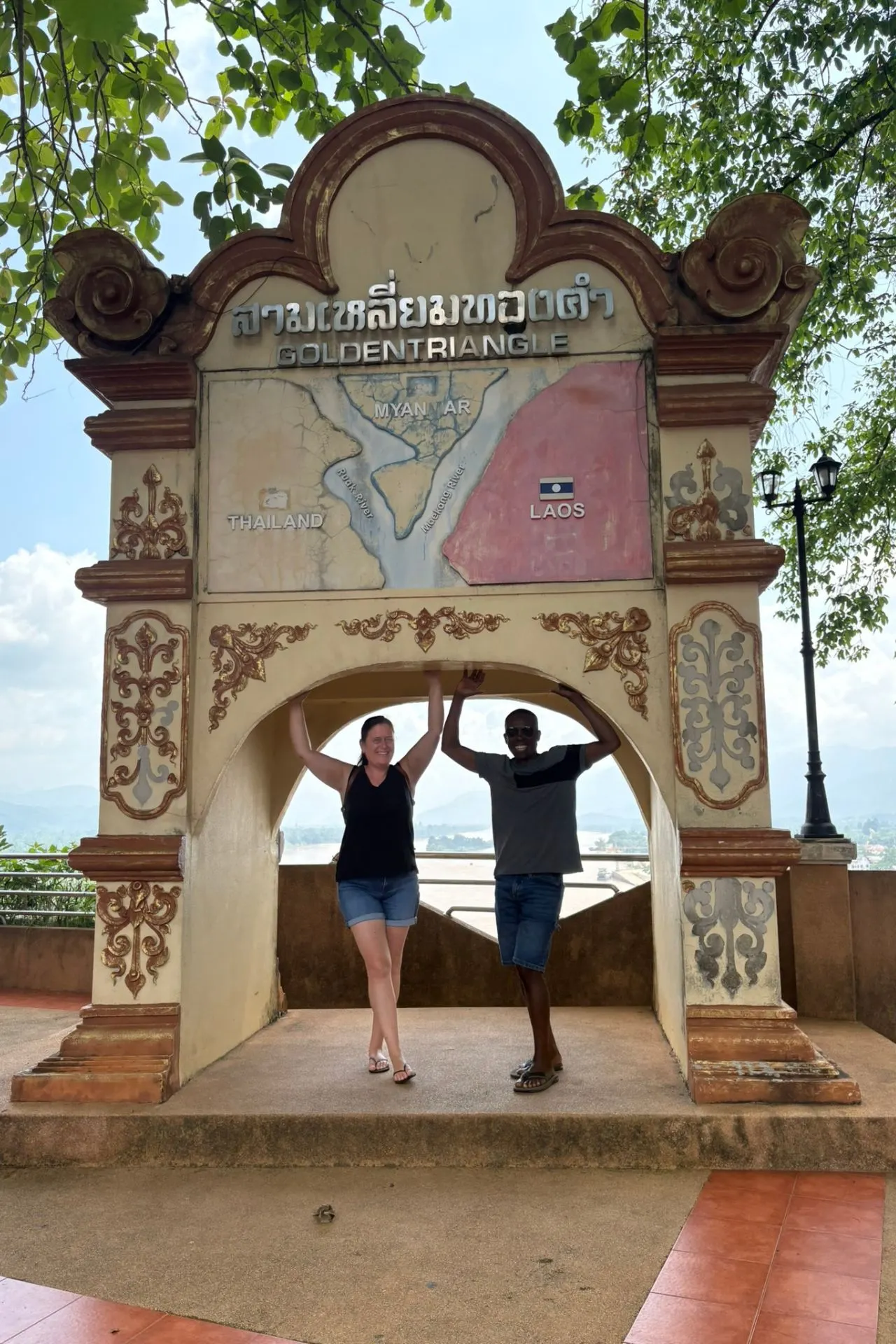 Twee mensen poseren lachend onder een boog met de kaart van de Gouden Driehoek, waar Thailand, Myanmar en Laos samenkomen.