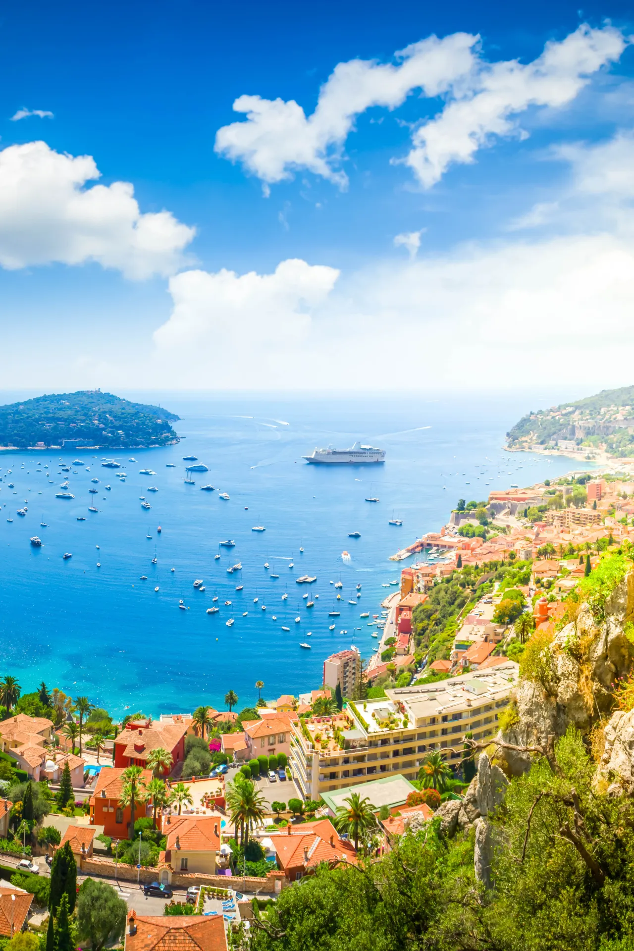 Panoramisch uitzicht op een kustplaats met rode daken en groene vegetatie die uitkijkt over een helderblauwe baai vol kleine boten en een groot cruiseschip in de verte. De kustlijn kronkelt langs heuvels en rotsen, terwijl op de achtergrond een eiland zichtbaar is onder een strakblauwe lucht met enkele witte wolken. Deze foto straalt een mediterrane sfeer uit, ideaal voor luxe vakanties, cruises en het ontdekken van schilderachtige kustbestemmingen.