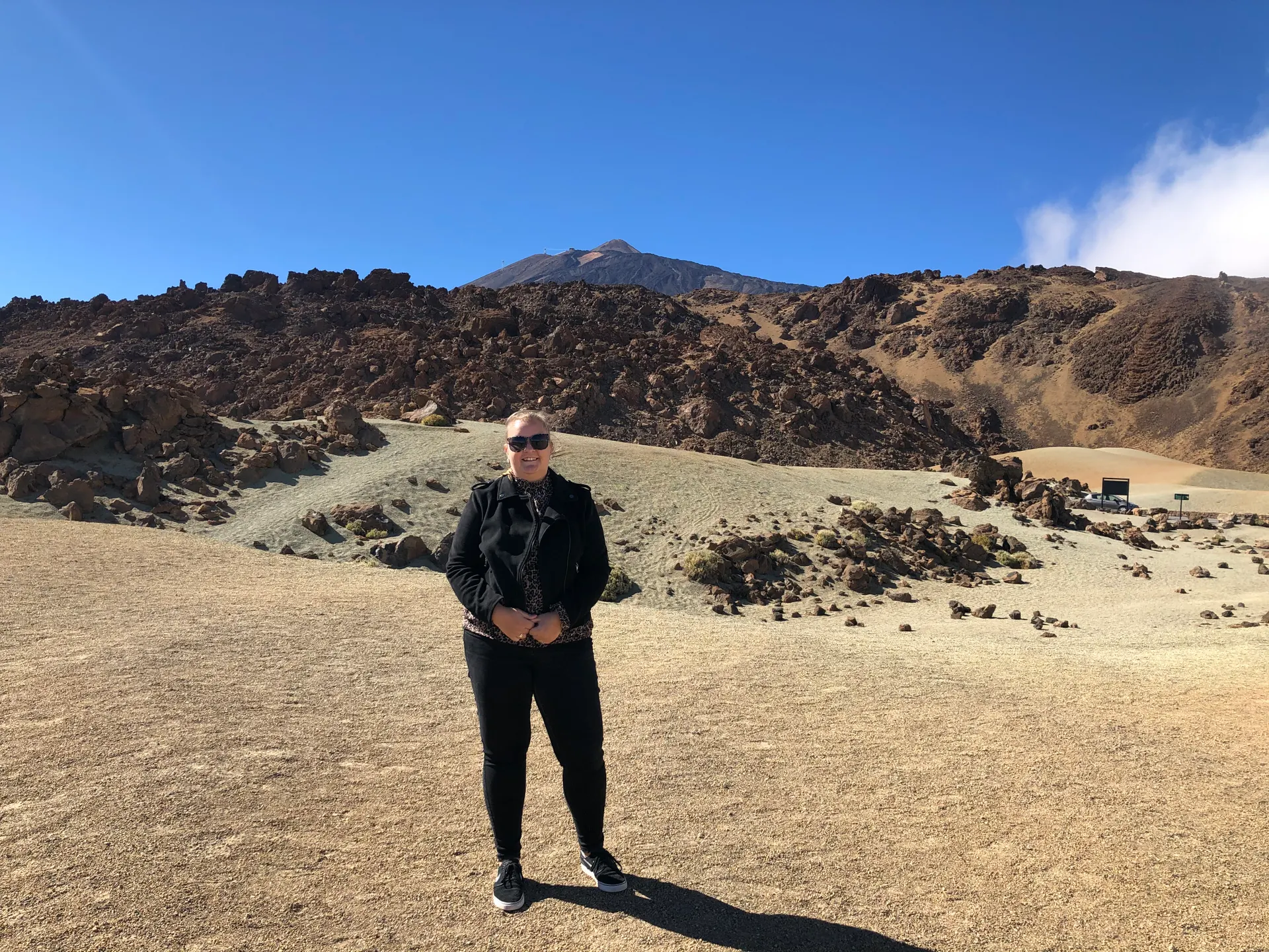 Foto in El Teide, Tenerife. Dame in het zwart gekleed met een zonnebril op staat in het midden van de foto tussen een bruin gebergte met een heldere blauwe lucht.