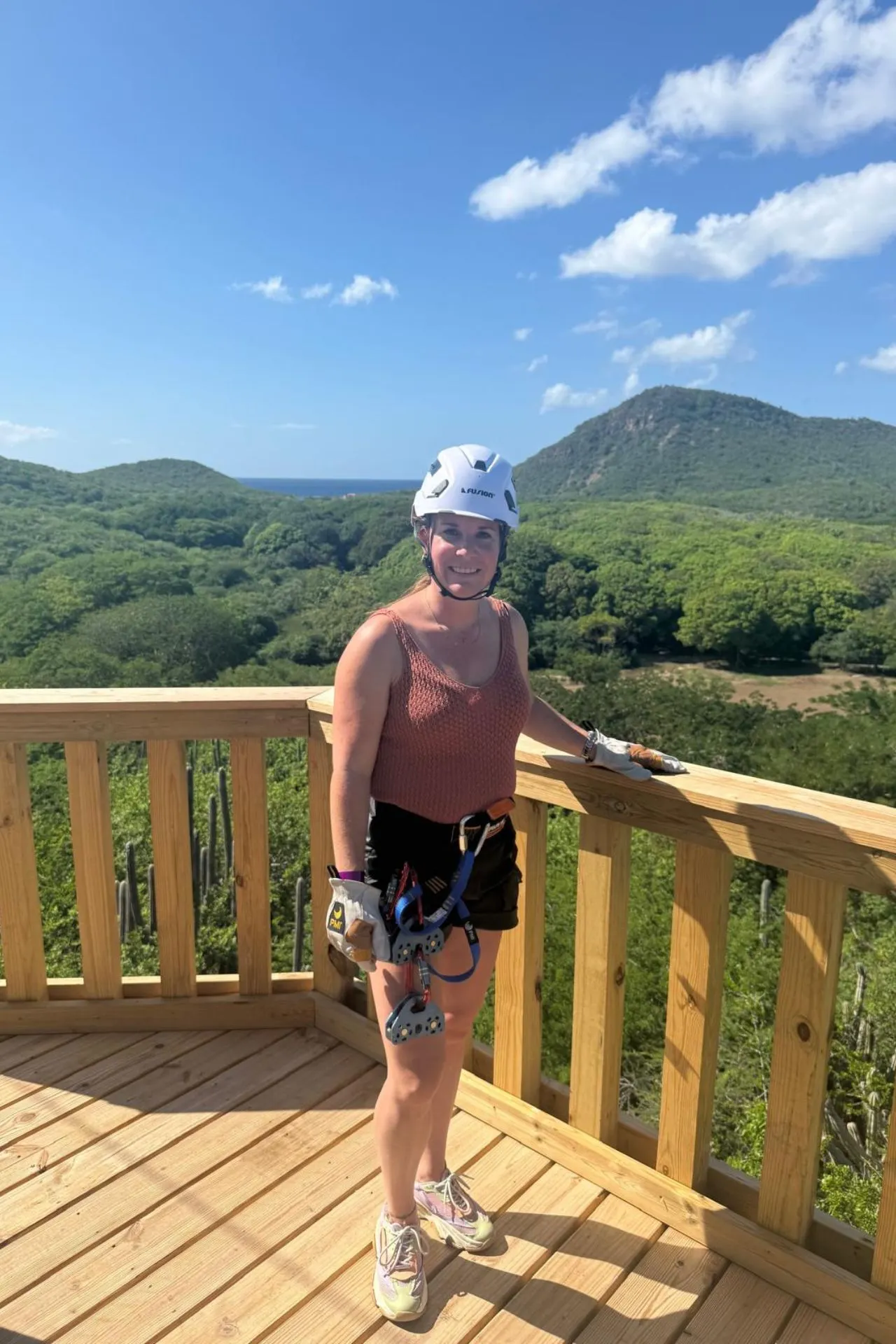 Persoon staat op een houten uitkijkplatform in een natuurgebied op Curaçao, uitgerust voor zipline of klimactiviteit. Op de achtergrond groene heuvels, cactussen en een glimp van de blauwe zee. Ideaal voor avontuurlijke reizigers die houden van outdoor activiteiten, natuur en adrenaline tijdens een vakantie in het Caribisch gebied.