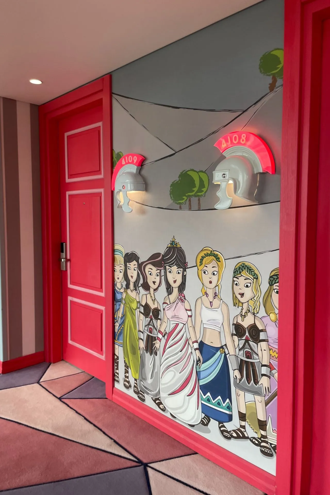 Rode deur van een hotelkamer met een striptekening van prinsessenfiguren en twee lampen in de vorm van olifanten.
