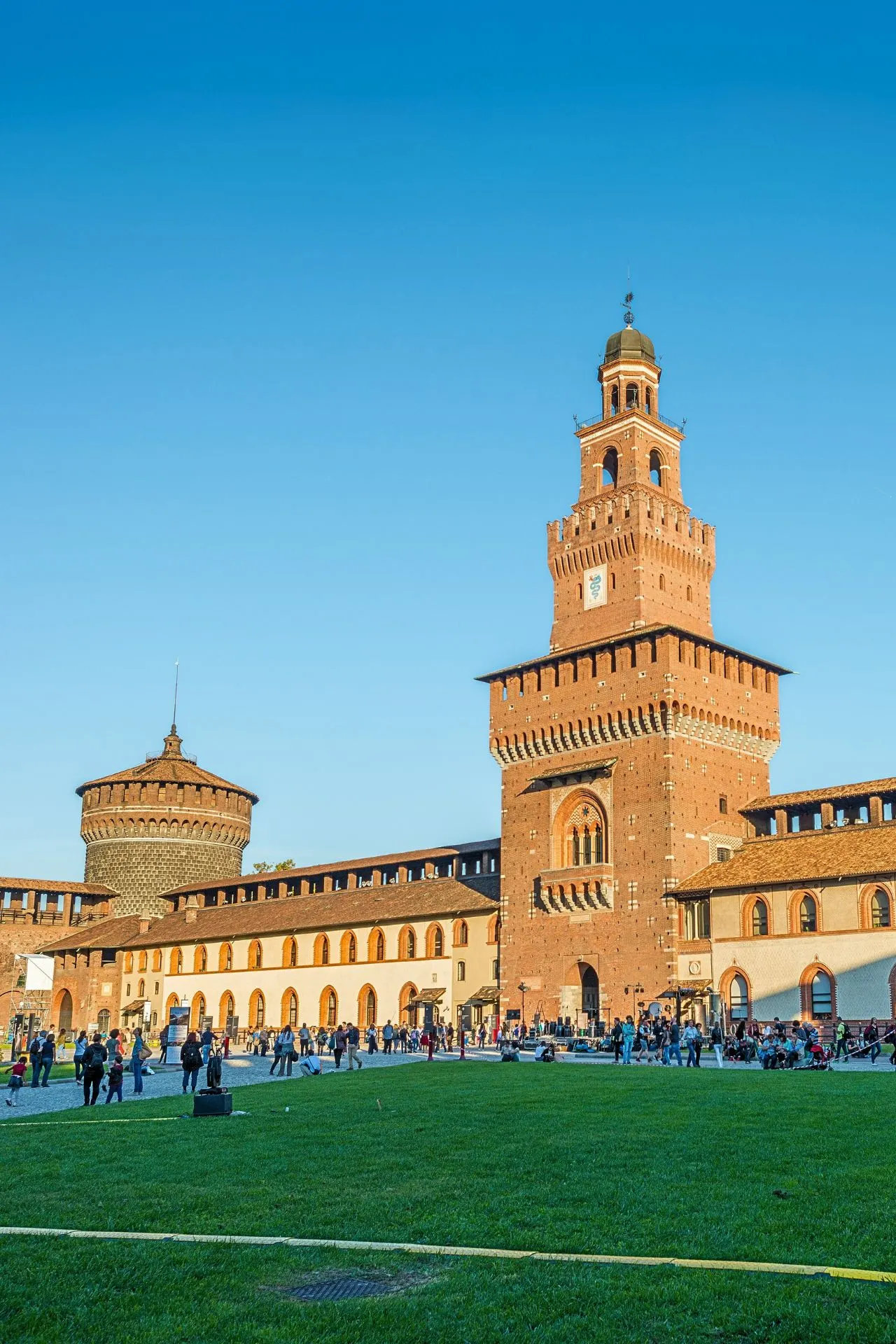 Het Castello Sforzesco in Milaan, een imposant middeleeuws kasteel met hoge bakstenen torens en een levendig plein met bezoekers op het grasveld.