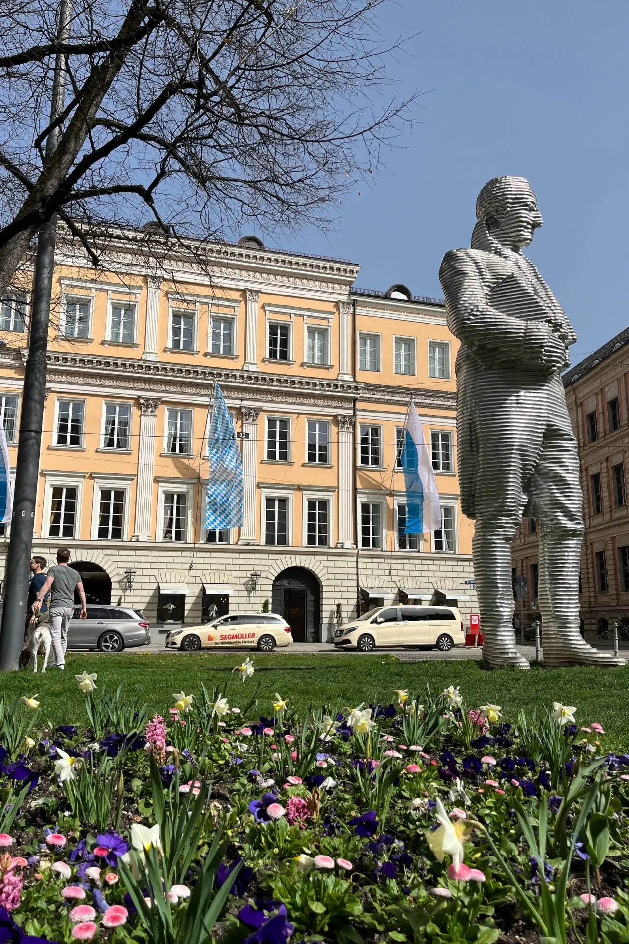 Een moderne sculptuur in een park in München, met bloeiende bloemen op de voorgrond en een klassieke architecturale achtergrond.