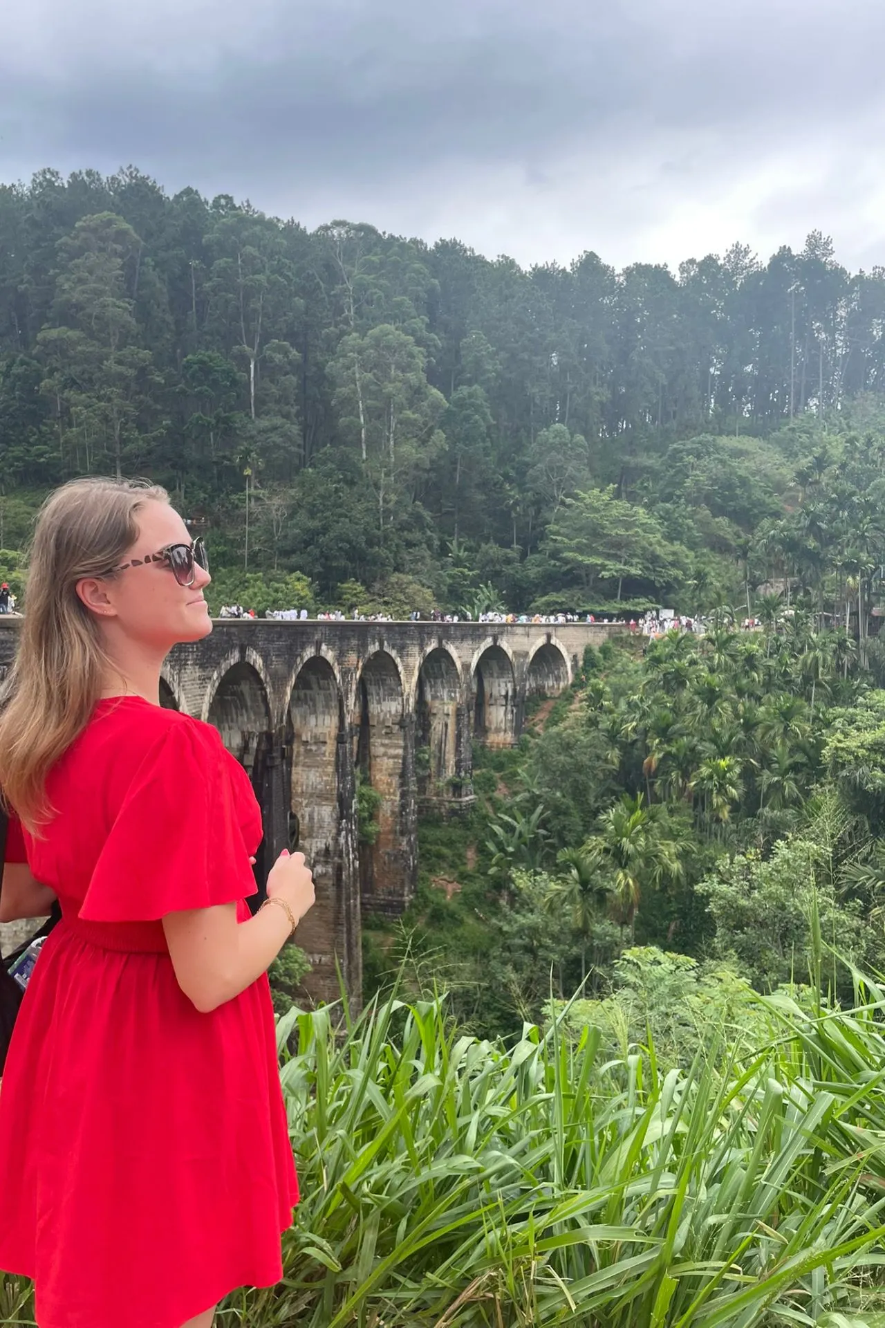 Een vrouw in een opvallende rode jurk poseert met uitzicht op de Nine Arches Bridge in Ella. Ze kijkt dromerig naar de brug waar andere reizigers wandelen, terwijl de groene jungle en hoge bomen het landschap dramatisch omarmen onder een bewolkte hemel. Een moment van bewondering en rust te midden van Sri Lanka’s natuurlijke pracht.