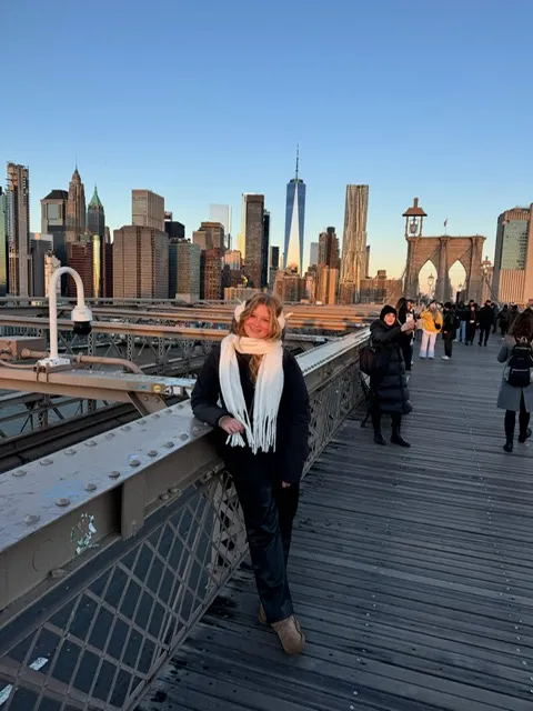 Een blonde vrouw staat op een brug met de indrukwekkende skyline van New York op de achtergrond, terwijl de zon de stad een warme gloed geeft.