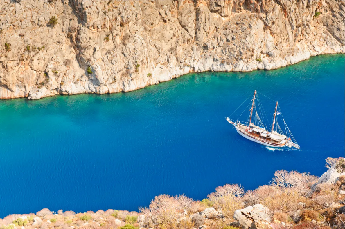 Traditioneel zeilschip vaart door een smalle baai met helderblauw water, omringd door indrukwekkende rotsformaties op een Grieks eiland. De steile kliffen contrasteren prachtig met het diepe blauw van de zee, terwijl het houten schip een authentieke en avontuurlijke sfeer toevoegt. Deze idyllische locatie is perfect voor een vakantie in Griekenland, met mogelijkheden voor boottochten, natuurbeleving en het ontdekken van verborgen kustlijnen.