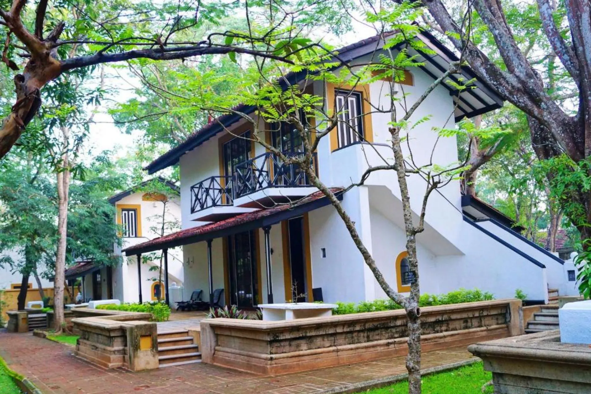 Witte villa in koloniale stijl bij Cinnamon Lodge Habarana met balkon en terras, omgeven door tropische bomen en rode paden door een weelderige tuin.


