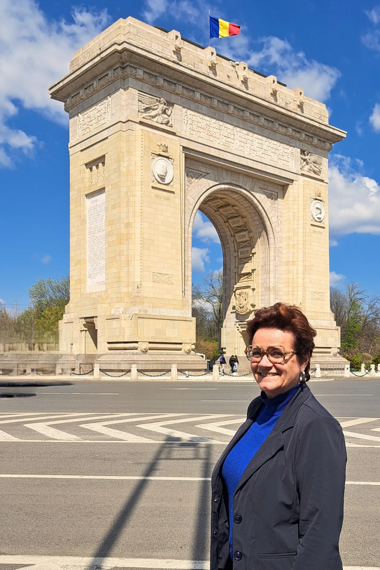 Vrouw met kort roodbruin haar en bril poseert voor een stenen boog met de Roemeense vlag op de achtergrond