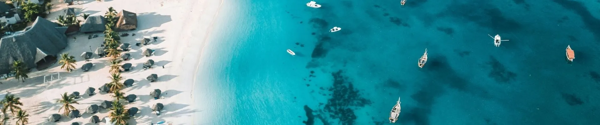Luchtfoto van Zanzibar met een hagelwit zandstrand, traditionele strandaccommodaties tussen palmbomen en het helderblauwe water van de Indische Oceaan. In de turquoise lagune liggen meerdere boten voor anker. Deze tropische kustlijn laat de paradijselijke stranden, rust en natuurlijke schoonheid van Zanzibar zien.