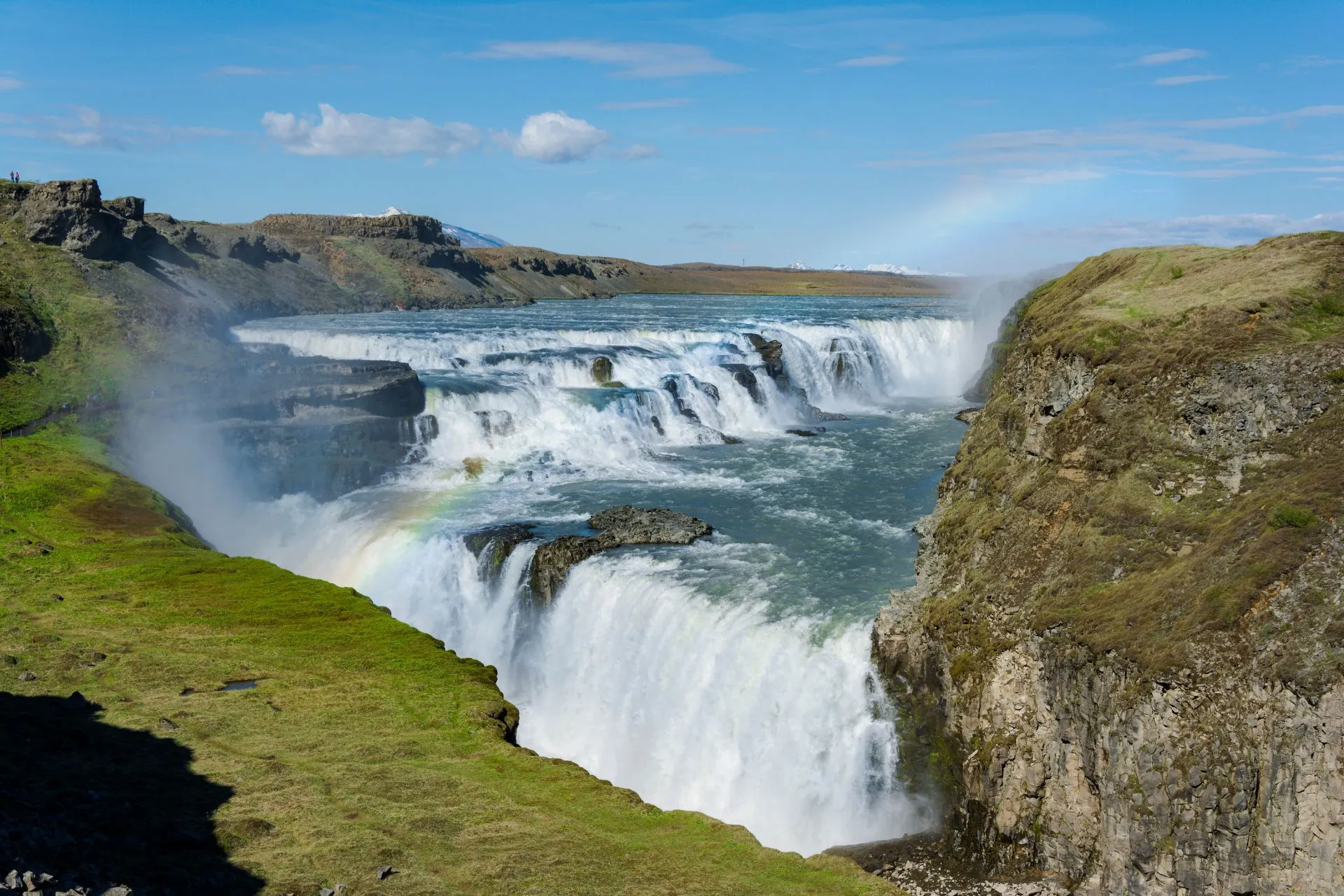 De krachtige Gullfoss waterval in IJsland met een regenboog in de nevel en omliggende groene en rotsachtige heuvels.