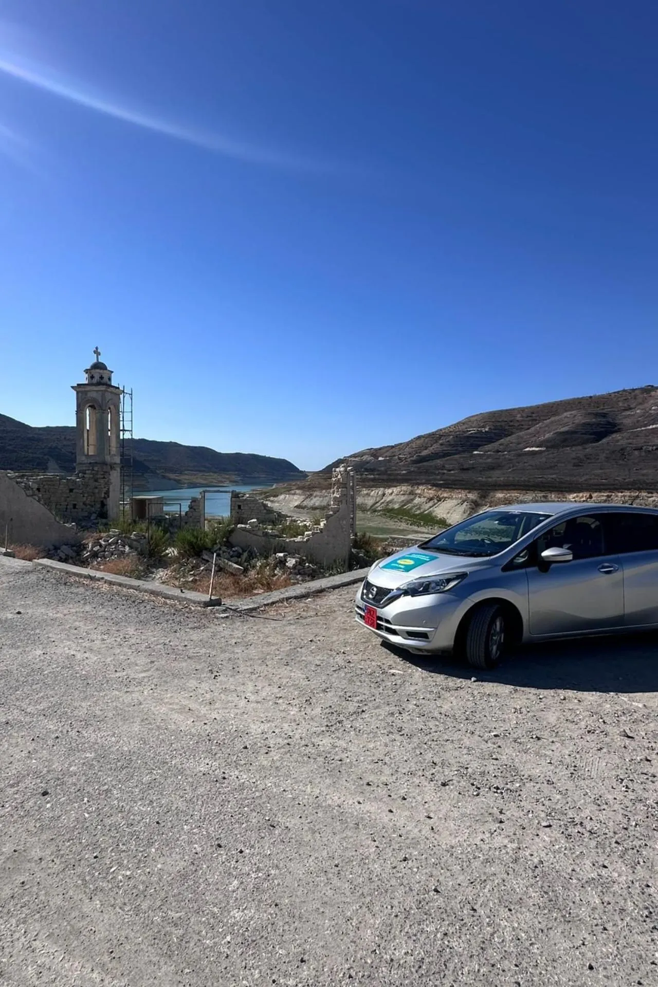 Witte huurauto van Sunny Cars geparkeerd op een stoffige weg in Cyprus, met een oude torenruïne en een meer in de achtergrond.