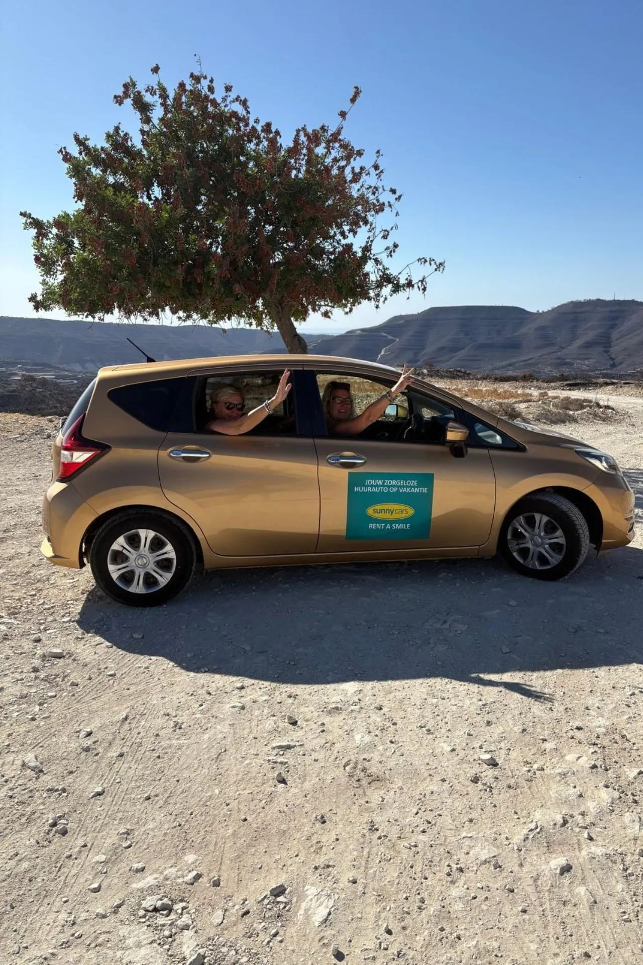 Gouden huurauto van Sunny Cars op een bergachtige, stoffige weg in Cyprus, met een boom en heuvels op de achtergrond. Twee personen zitten in de auto en steken hun armen uit het raam.