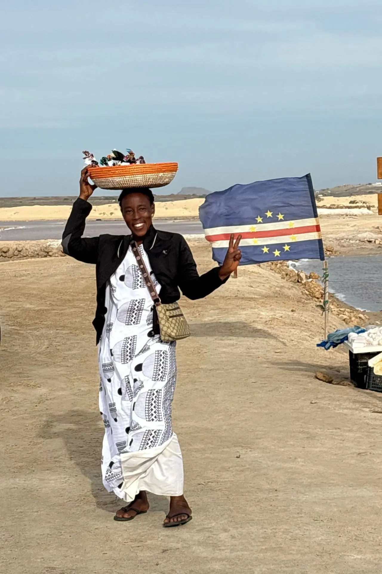 Een persoon staat buiten op een zandpad bij zoutvlaktes, houdt een kleurrijke mand op het hoofd en maakt een vreedzaam handgebaar terwijl een Kaapverdische vlag wappert naast hen.