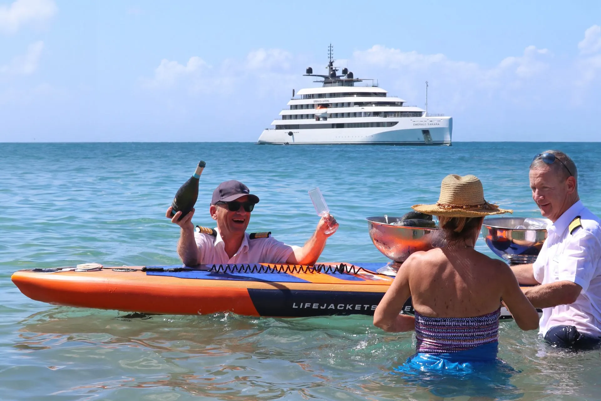 Een ontspannen strandervaring waarin gasten genieten van champagne in het heldere ondiepe water, met op de achtergrond een stijlvol superjacht dat voor de kust ligt. De setting straalt exclusiviteit en persoonlijke service uit, typerend voor een luxe cruise all inclusive waarin elke activiteit met zorg wordt verzorgd. De tropische omgeving en turquoise zee versterken het gevoel van ultiem vakantiegenot tijdens een luxe cruise vakantie.