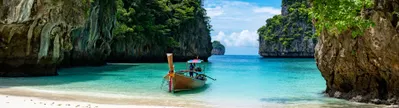 Een serene, wit zandstrand in Thailand met helderblauw water en een enkele, traditionele houten boot die in het midden van het water drijft. Omringende rotsformaties reiken hoog omhoog en laten een doorkijk naar de open zee in het midden, waar het zonlicht schittert op het kristalheldere water.
