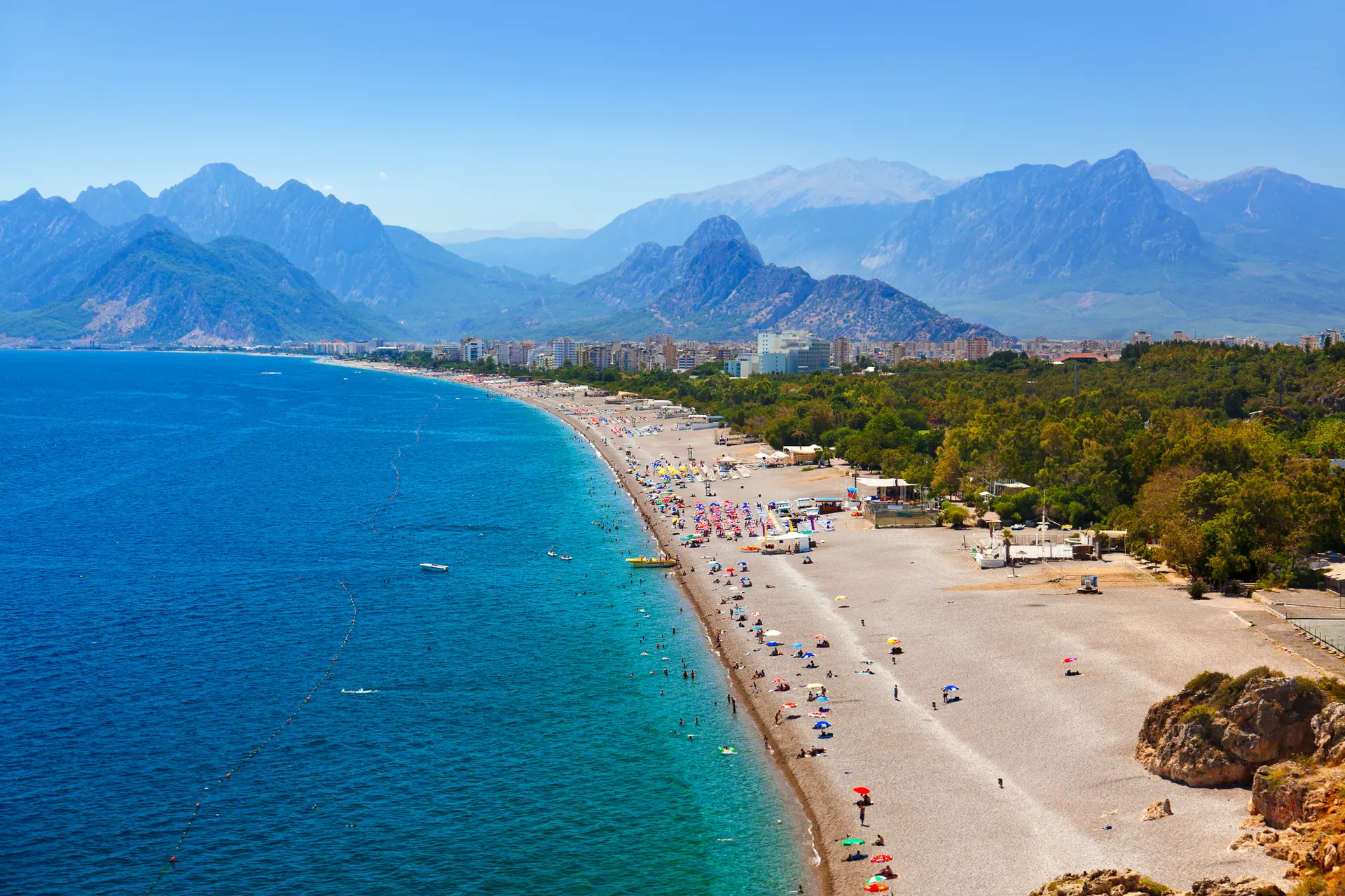 “Panoramisch uitzicht op het Konyaalti-strand in Antalya, Turkije, met een lange kustlijn langs helderblauw water van de Middellandse Zee en groene bergen op de achtergrond onder een stralend blauwe lucht. Het strand is bezaaid met parasols en badgasten, ideaal voor zonvakantie, all-inclusive resorts en zomervakantie in Turkije. Perfecte bestemming voor ontspanning, strandactiviteiten en het ontdekken van de Turkse Rivièra.”